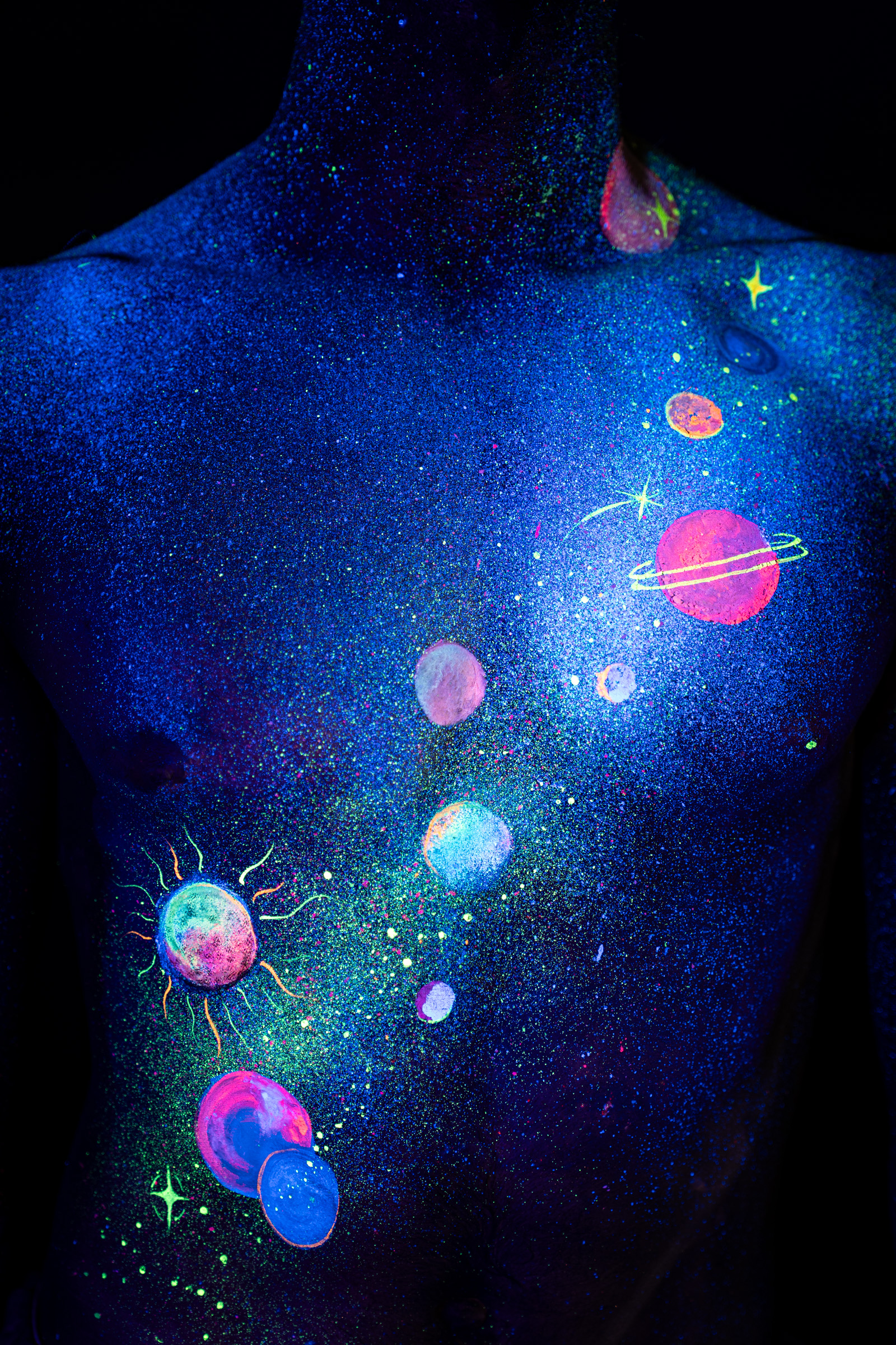 Axell, standing and facing the camera. The same space themed design of glowing pink, orange and blue planets continues down the chest and abdomen over a glittering blue base. Bodypaint by Elad Itzkin, Charlotte Bobineau, Sage Brewer. Photographed by Elad Itzkin Photography under UV/blacklight in his studio in Gesundbrunnen, Berlin.