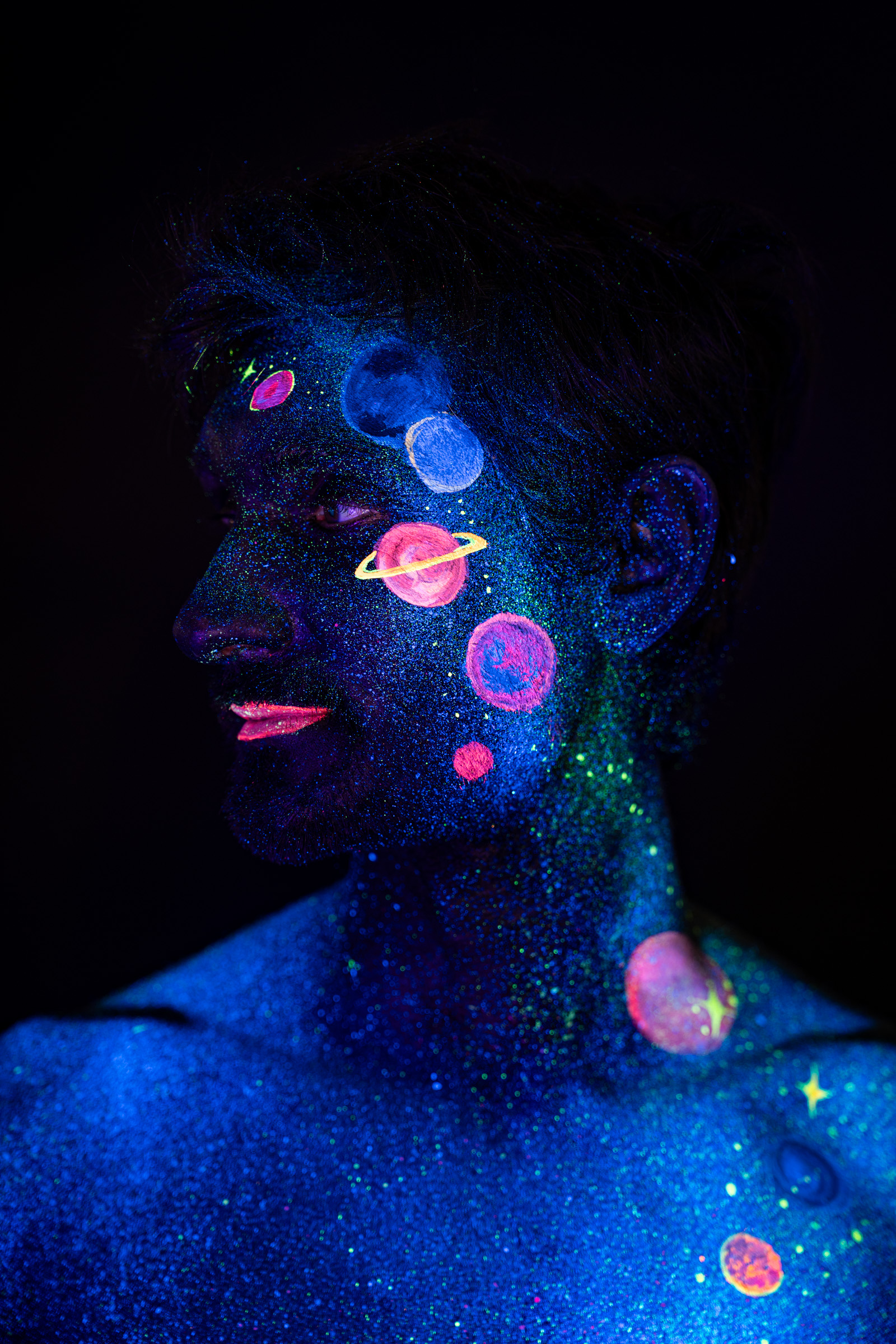 Axell, in a three-quarter profile view of the head and shoulders. Glowing pink, orange and blue planets form a space themed design across the face and neck. Bodypaint by Elad Itzkin, Charlotte Bobineau, Sage Brewer. Photographed by Elad Itzkin Photography under UV/blacklight in his studio in Gesundbrunnen, Berlin.