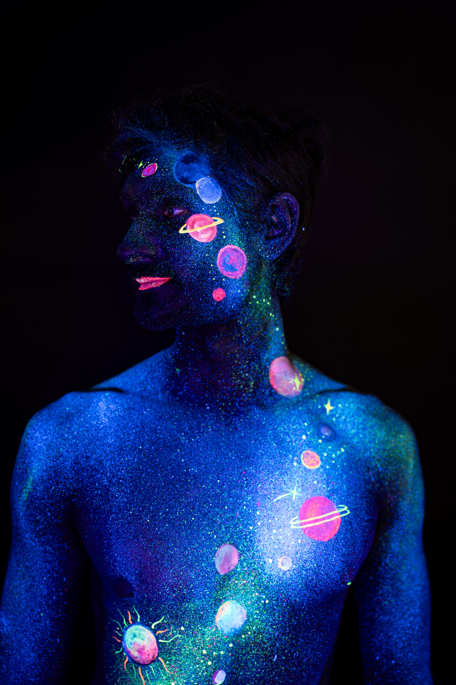 Axell, in a three-quarter profile view of the upper body. A space themed design of glowing pink, orange and blue planets spans the face, neck and chest. Bodypaint by Elad Itzkin, Charlotte Bobineau, Sage Brewer. Photographed by Elad Itzkin Photography under UV/blacklight in his studio in Gesundbrunnen, Berlin.