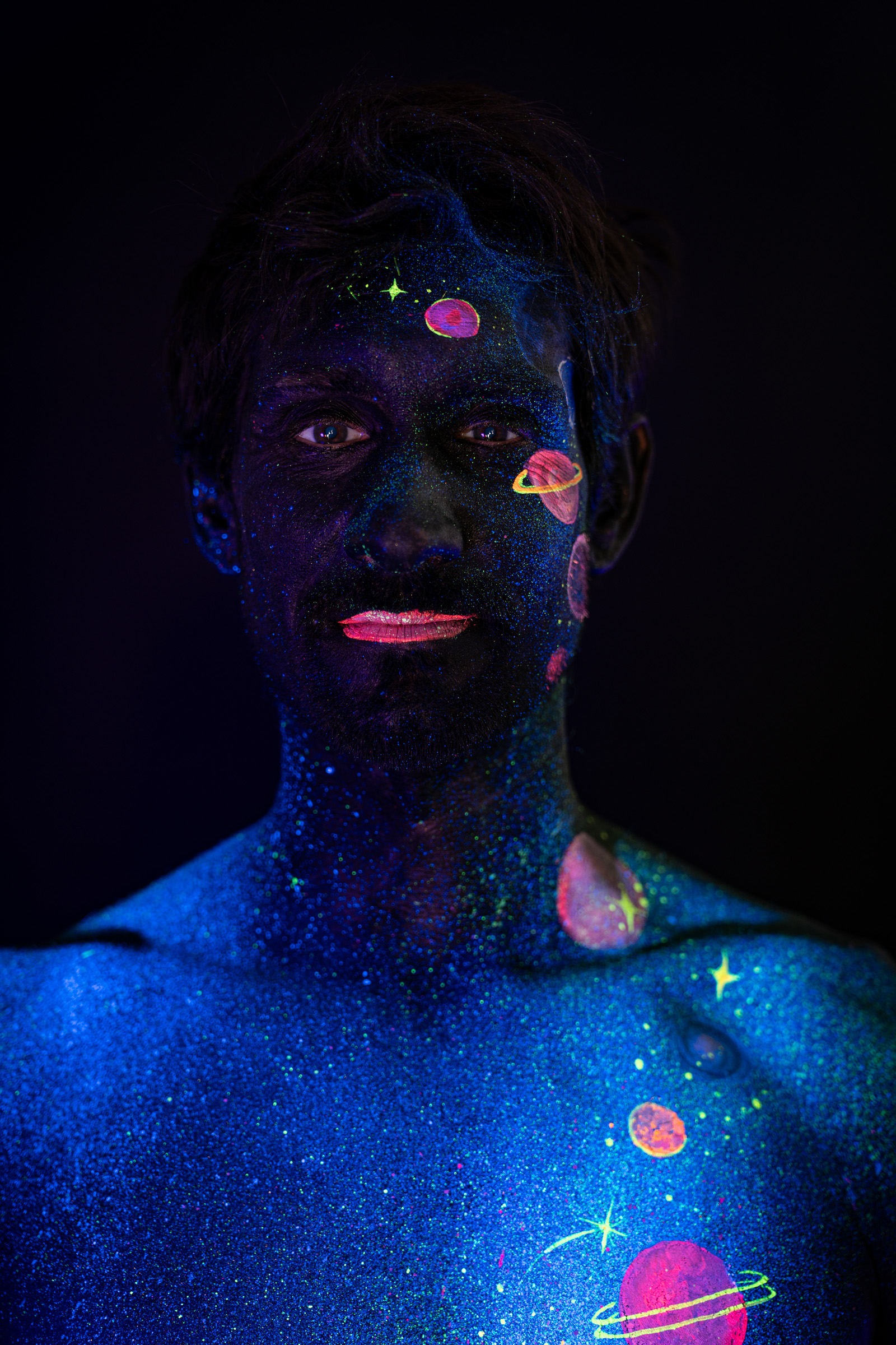 Axell, standing and facing the camera directly. Glowing pink, orange and blue planets form a space themed design across the face and chest over a glittering blue base. Bodypaint by Elad Itzkin, Charlotte Bobineau, Sage Brewer. Photographed by Elad Itzkin Photography under UV/blacklight in his studio in Gesundbrunnen, Berlin.