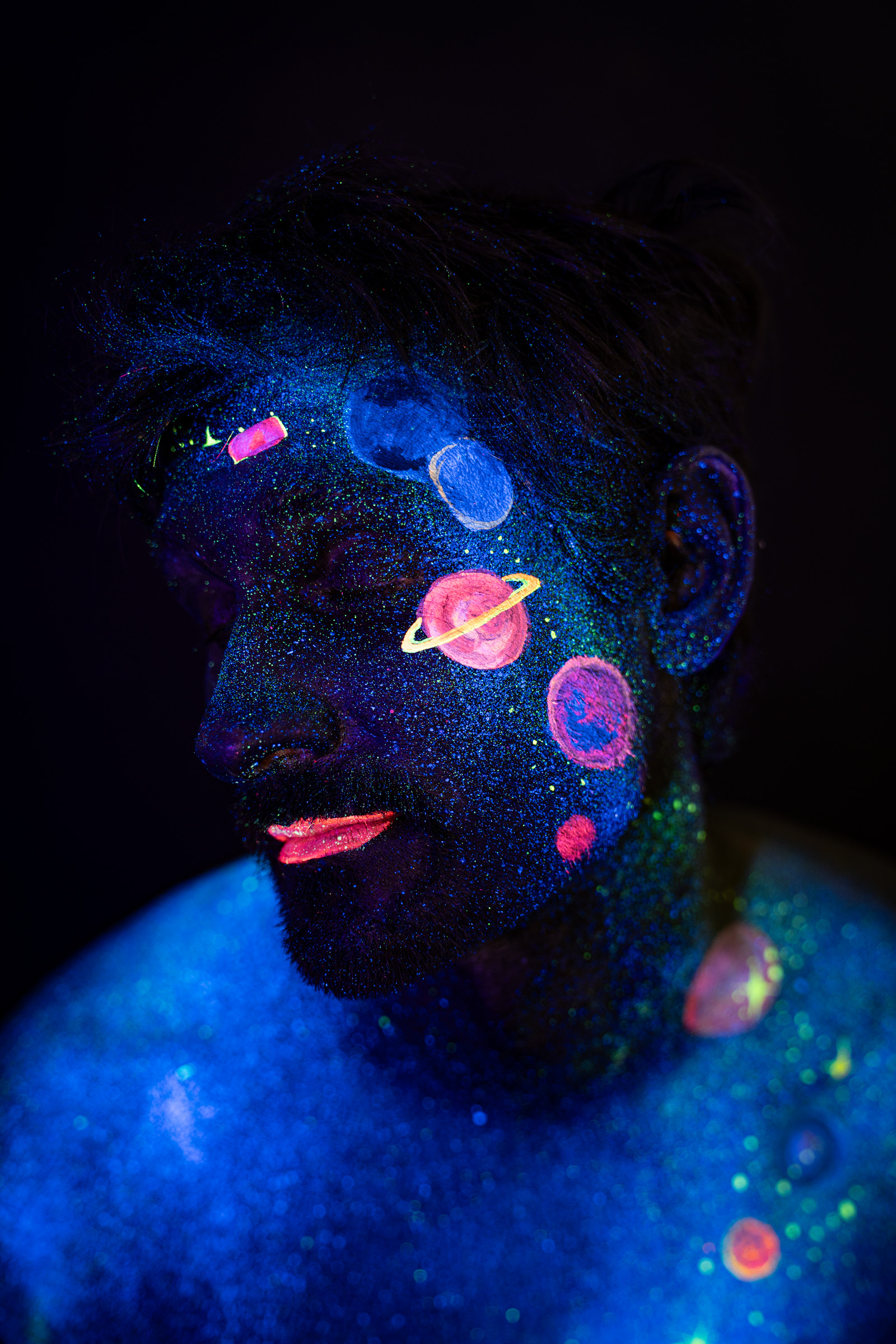 Axell, in a close three-quarter profile view, a variation of the same pose. Glowing pink, orange and blue planets form a space themed design across the face. Bodypaint by Elad Itzkin, Charlotte Bobineau, Sage Brewer. Photographed by Elad Itzkin Photography under UV/blacklight in his studio in Gesundbrunnen, Berlin.