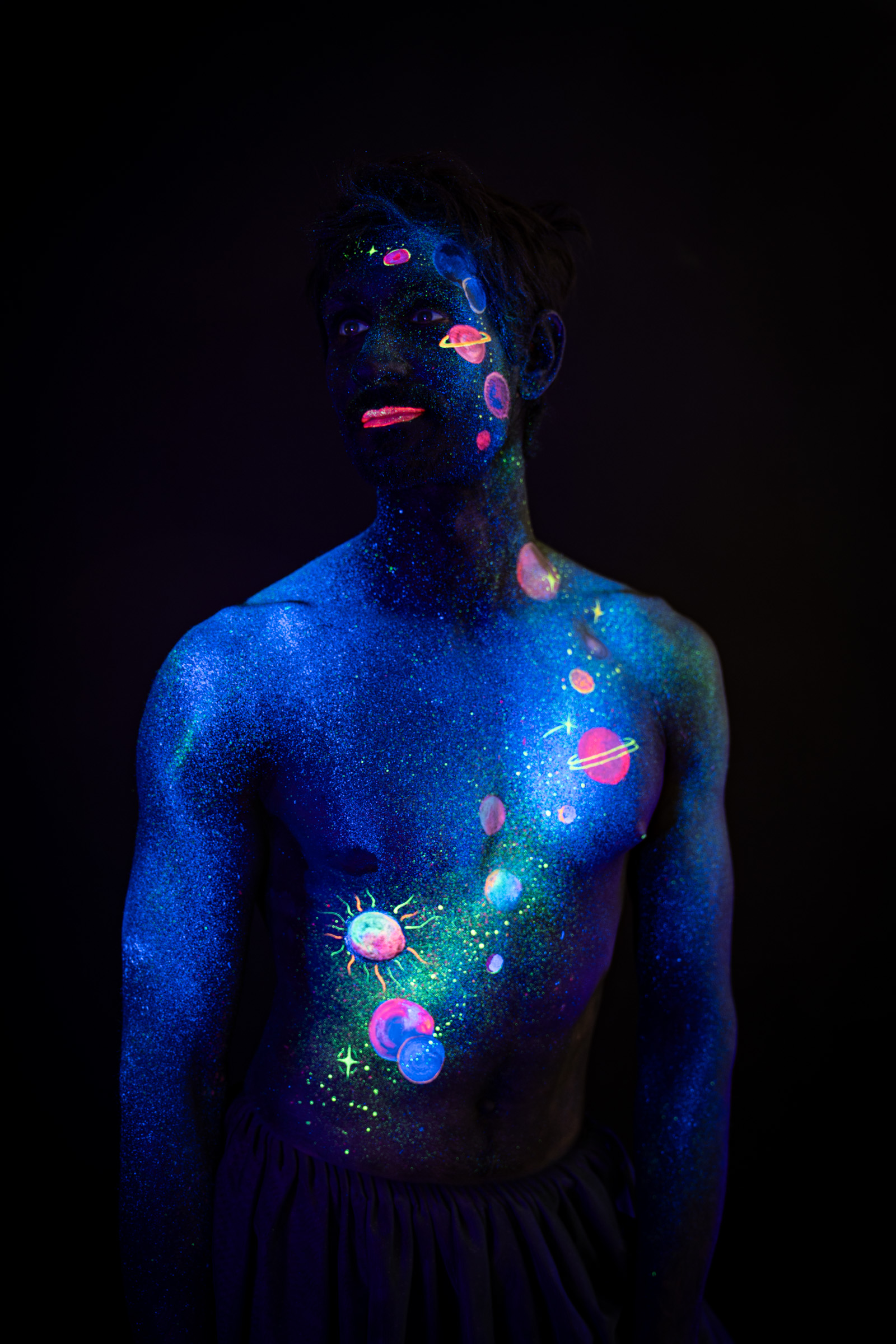 Axell, standing and facing forward with the upper body in view. A vertical row of glowing pink, orange and blue planets runs down the center of the torso. Bodypaint by Elad Itzkin, Charlotte Bobineau, Sage Brewer. Photographed by Elad Itzkin Photography under UV/blacklight in his studio in Gesundbrunnen, Berlin.
