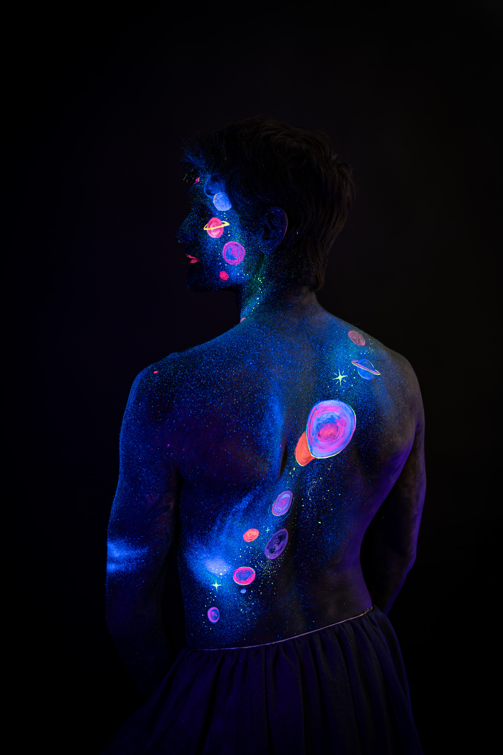 Axell, standing with the back turned toward the camera, the head turned to show the face in profile. Glowing pink, orange and blue planets line the back. Bodypaint by Elad Itzkin, Charlotte Bobineau, Sage Brewer. Photographed by Elad Itzkin Photography under UV/blacklight in his studio in Gesundbrunnen, Berlin.