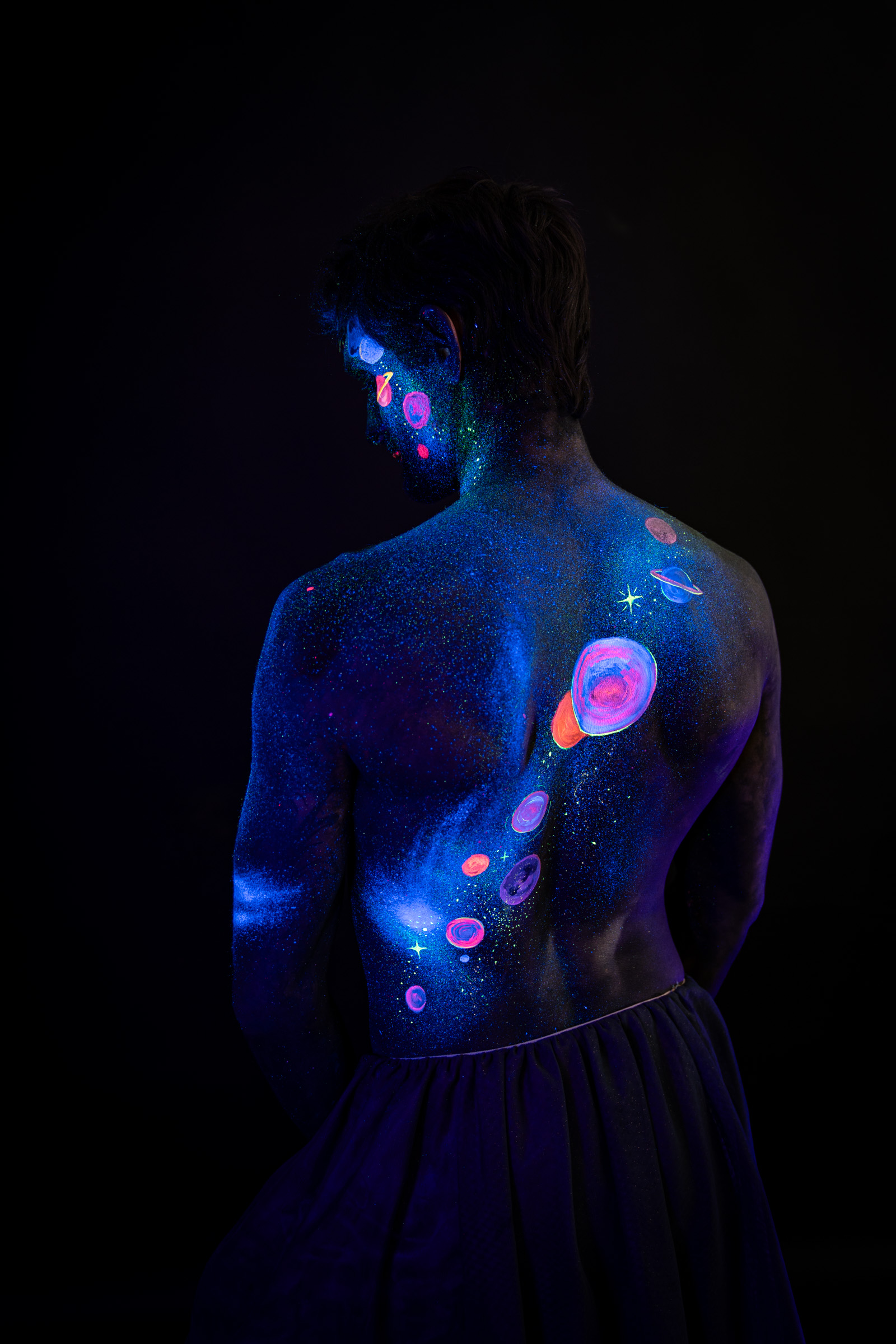 Axell, standing with the back to the camera, a variation of the same three-quarter pose with the face in profile. Glowing planets in pink, orange and blue run down the back. Bodypaint by Elad Itzkin, Charlotte Bobineau, Sage Brewer. Photographed by Elad Itzkin Photography under UV/blacklight in his studio in Gesundbrunnen, Berlin.