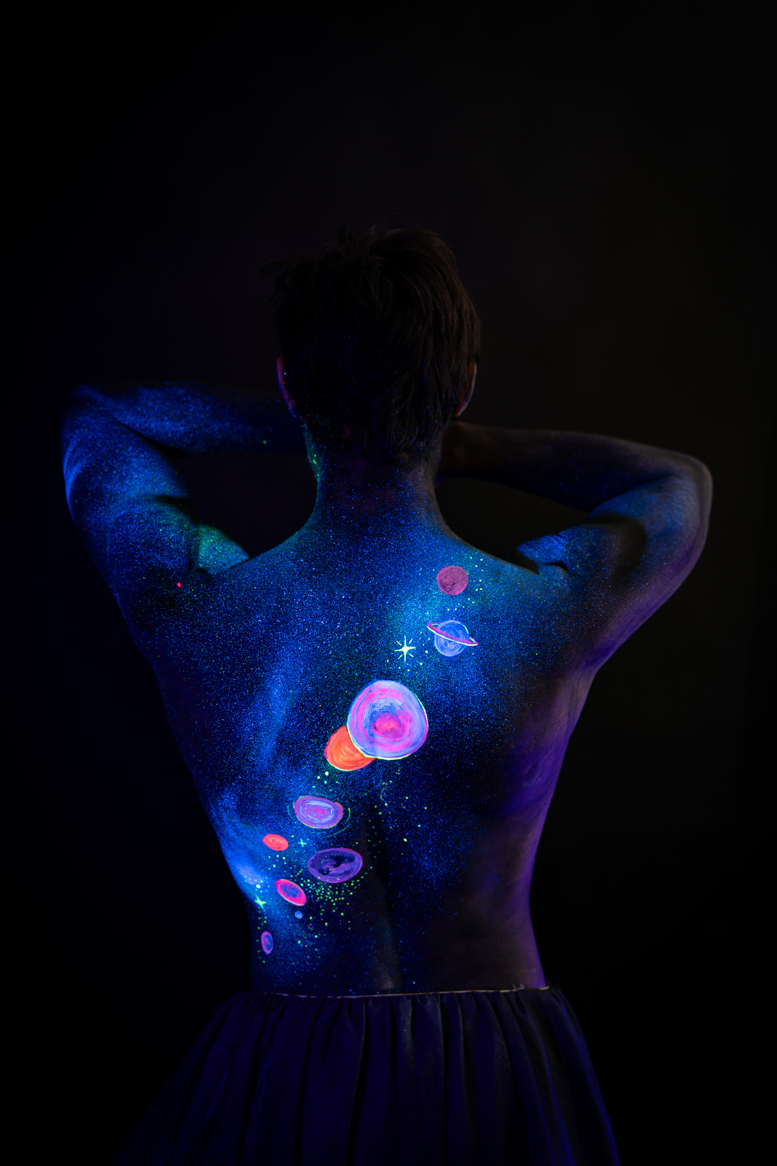 Axell, standing with the back fully turned to the camera. A space themed design of glowing pink, orange and blue planets runs down the upper back and spine. Bodypaint by Elad Itzkin, Charlotte Bobineau, Sage Brewer. Photographed by Elad Itzkin Photography under UV/blacklight in his studio in Gesundbrunnen, Berlin.