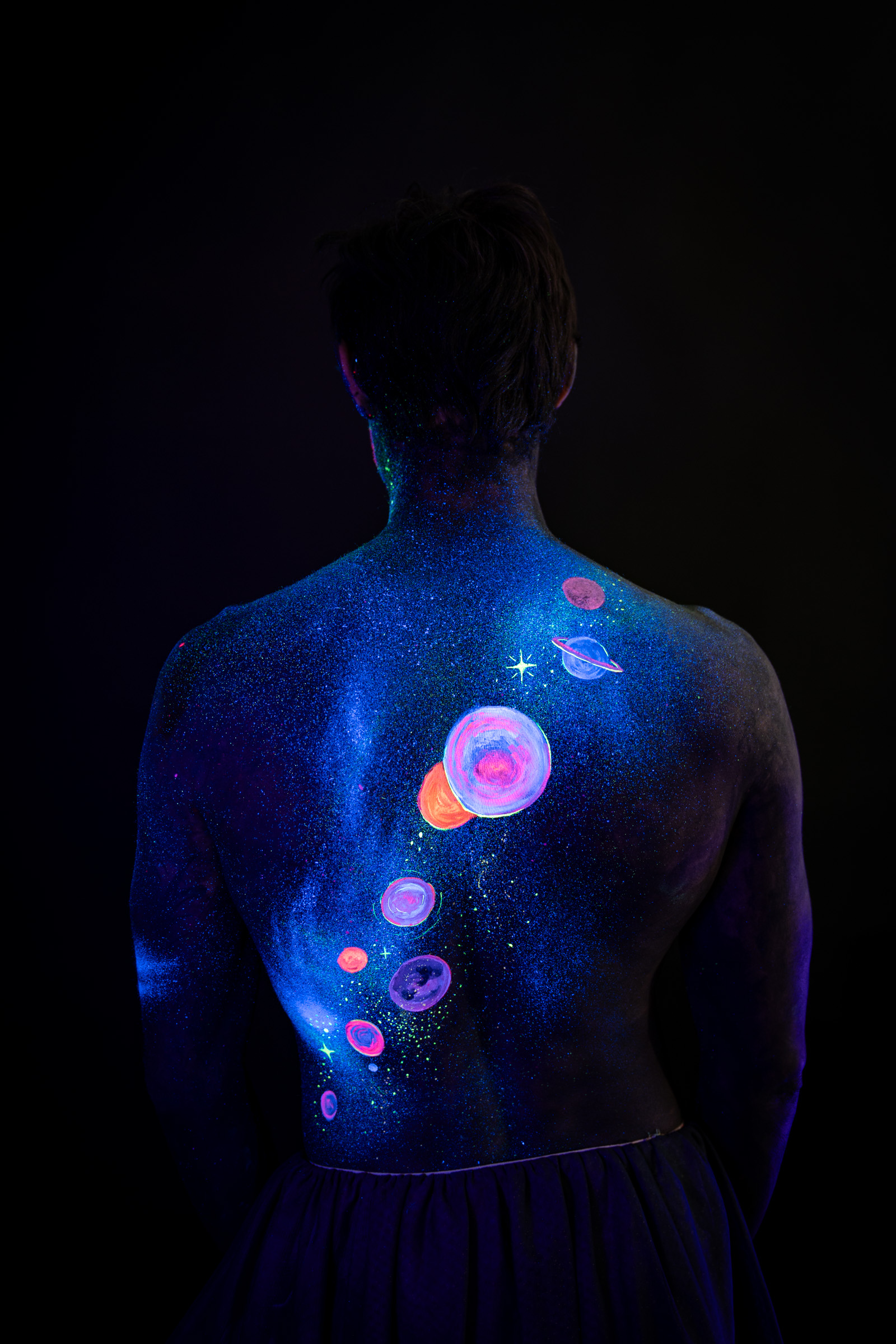 Axell, standing with the back squarely to the camera. A space themed design of glowing pink, orange and blue planets runs down the spine over a glittering blue base. Bodypaint by Elad Itzkin, Charlotte Bobineau, Sage Brewer. Photographed by Elad Itzkin Photography under UV/blacklight in his studio in Gesundbrunnen, Berlin.