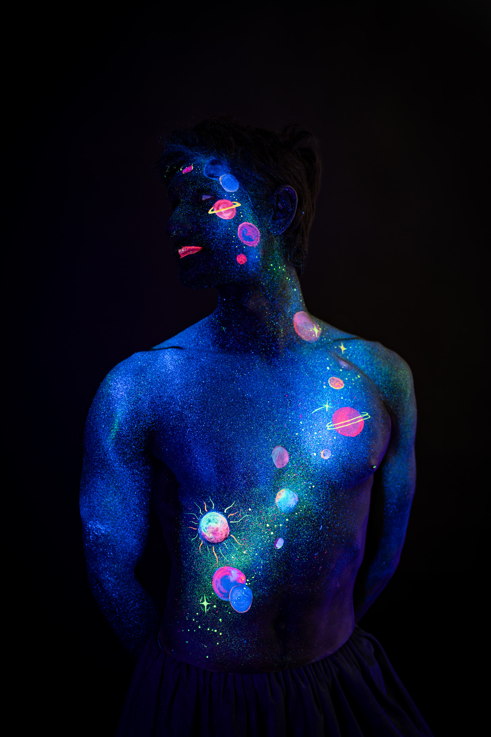 Axell, standing in a three-quarter side view, the upper body turned toward the camera. Glowing pink, orange and blue planets form a space themed design across the face and torso. Bodypaint by Elad Itzkin, Charlotte Bobineau, Sage Brewer. Photographed by Elad Itzkin Photography under UV/blacklight in his studio in Gesundbrunnen, Berlin.