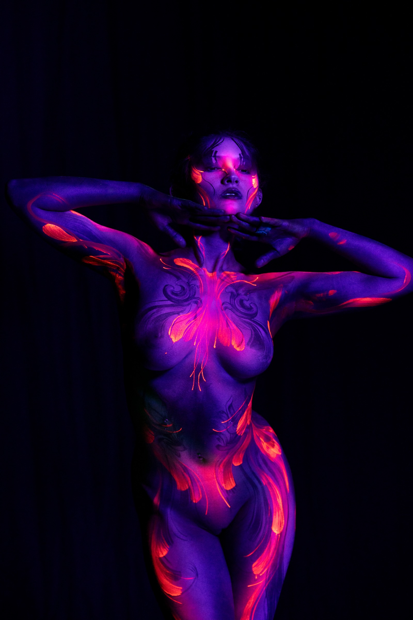 BabeyGianna stands with her head tilted back, her chest and torso covered in a butterfly-shaped orange and pink floral pattern over deep purple UV-reactive skin. Bodypaint by Claudia Kraemer, photographed by Elad Itzkin Photography under UV light in his studio in Gesundbrunnen, Berlin.