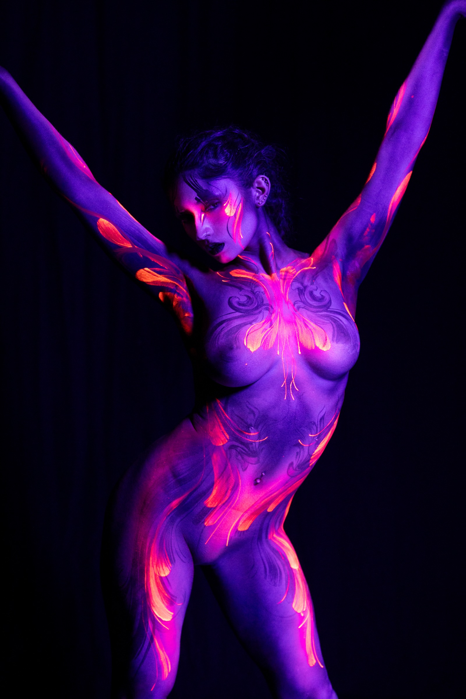BabeyGianna stands facing the camera, her chest, abdomen, and legs decorated with a symmetrical butterfly-shaped orange and pink floral pattern set against purple UV-lit skin. Bodypaint by Claudia Kraemer, photographed by Elad Itzkin Photography under UV light in his studio in Gesundbrunnen, Berlin.