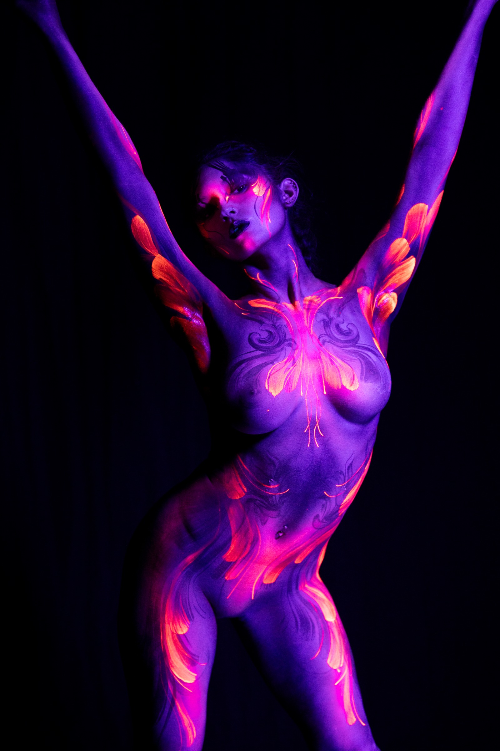 BabeyGianna stands facing forward, her torso painted with a butterfly-shaped orange and pink floral pattern that flows into flame-like linework across her purple UV-reactive skin. Bodypaint by Claudia Kraemer, photographed by Elad Itzkin Photography under UV light in his studio in Gesundbrunnen, Berlin.