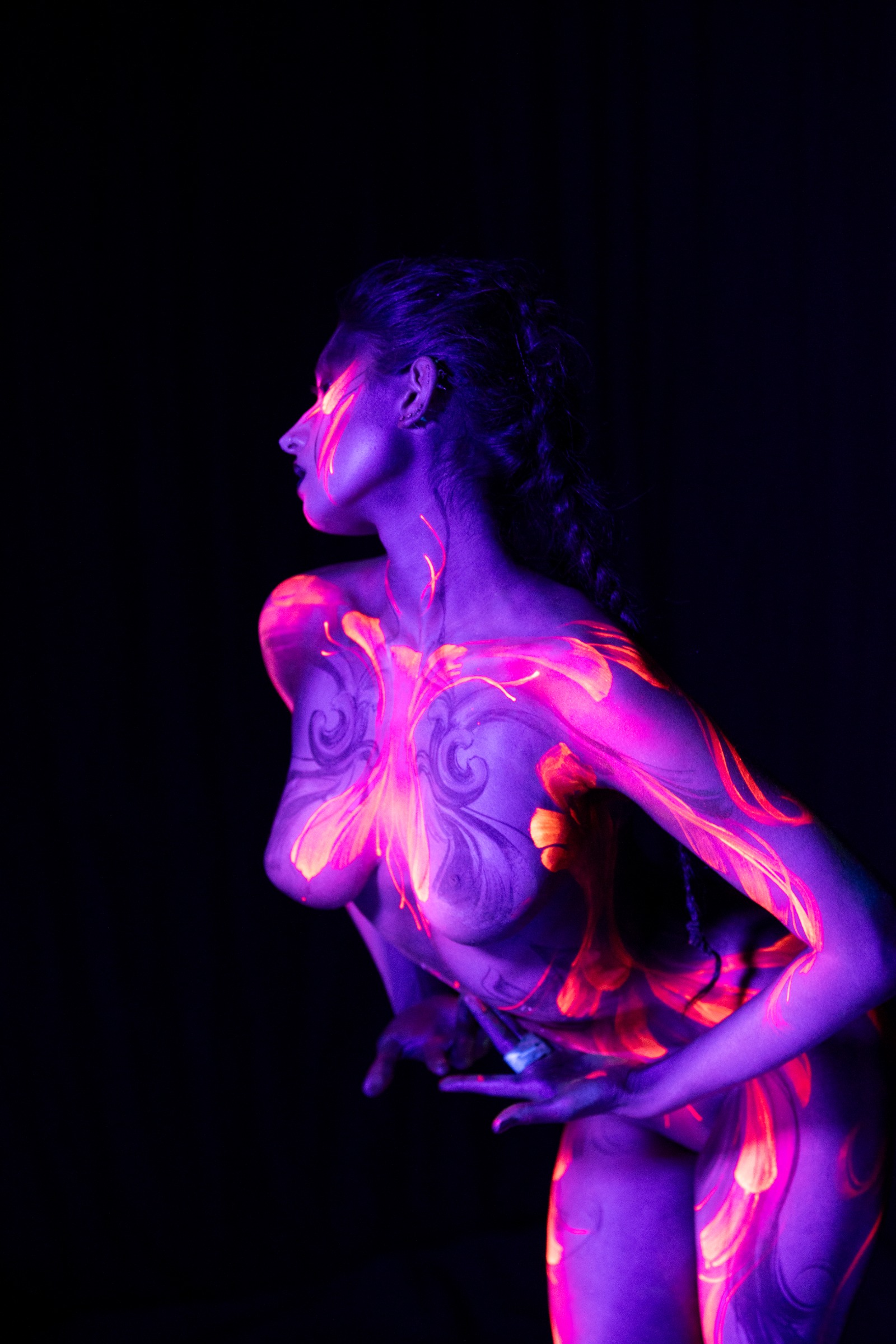 BabeyGianna stands in profile, bent slightly forward at the waist, her purple UV-lit torso and hip marked with a flowing orange and pink floral linework design. Bodypaint by Claudia Kraemer, photographed by Elad Itzkin Photography under UV light in his studio in Gesundbrunnen, Berlin.