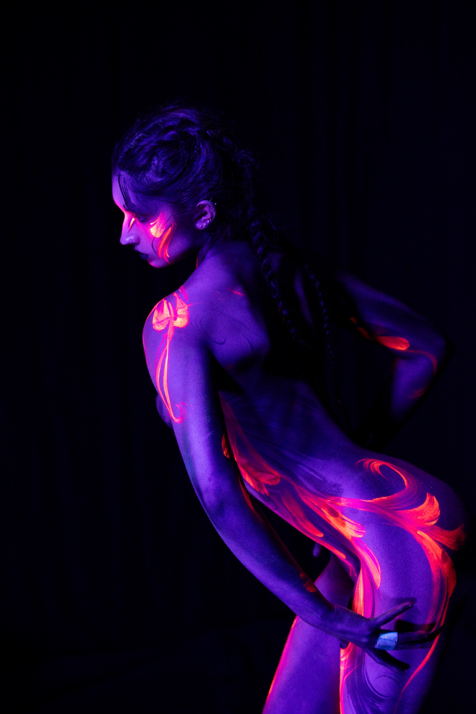 BabeyGianna stands in profile with her torso bent slightly forward, her braided hair visible as an orange and pink floral design sweeps across her purple UV-lit skin. Bodypaint by Claudia Kraemer, photographed by Elad Itzkin Photography under UV light in his studio in Gesundbrunnen, Berlin.