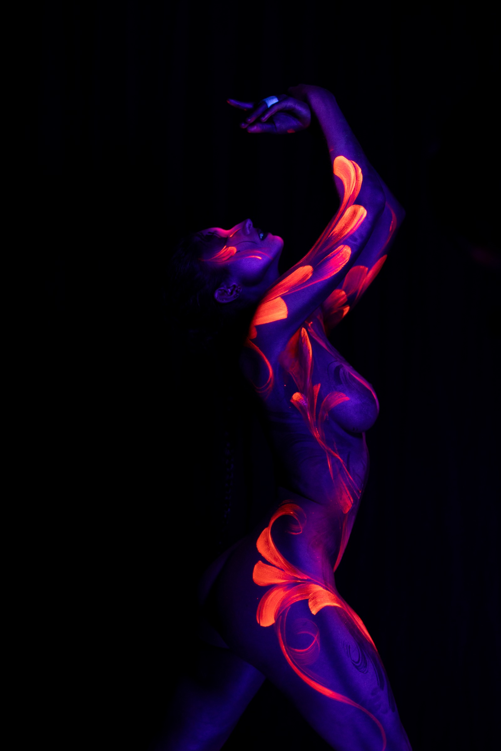 BabeyGianna stands with her torso turned to the side in a three-quarter view. Orange and pink floral linework spreads across a purple UV base on her torso and shoulder. Bodypaint by Claudia Kraemer. Photographed by Elad Itzkin Photography under UV light in Gesundbrunnen, Berlin.
