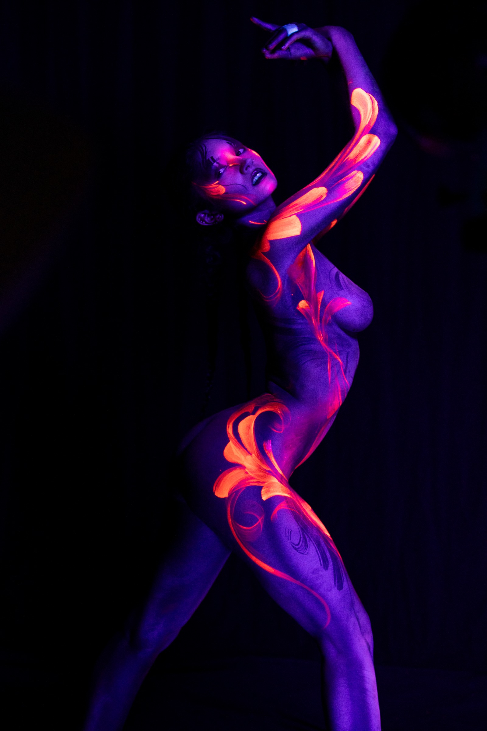 BabeyGianna stands with her torso in three-quarter view and her head tilted back. A violet UV base is patterned with orange and pink abstract floral linework across her chest and hip. Bodypaint by Claudia Kraemer. Photographed under blacklight by Elad Itzkin Photography in his Gesundbrunnen, Berlin studio.