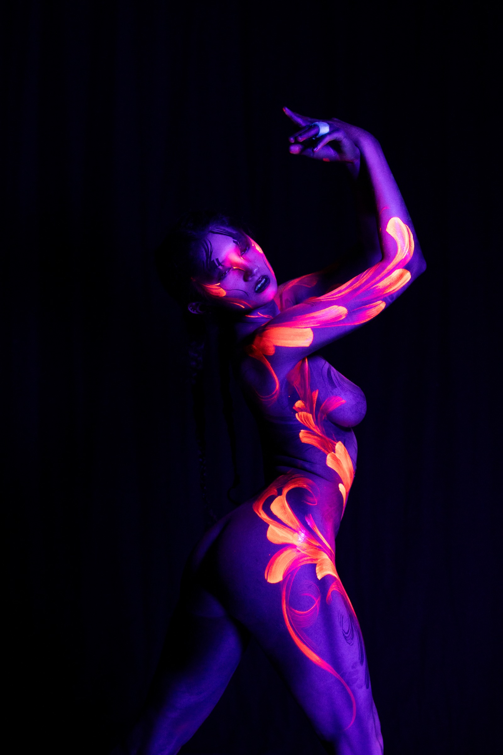 In a standing pose with her torso angled in three-quarter view, BabeyGianna's head is tilted back. Orange and pink floral linework covers a purple UV base across her chest, hip, and thigh. Bodypaint by Claudia Kraemer. Photographed by Elad Itzkin Photography under UV light in his Berlin studio.