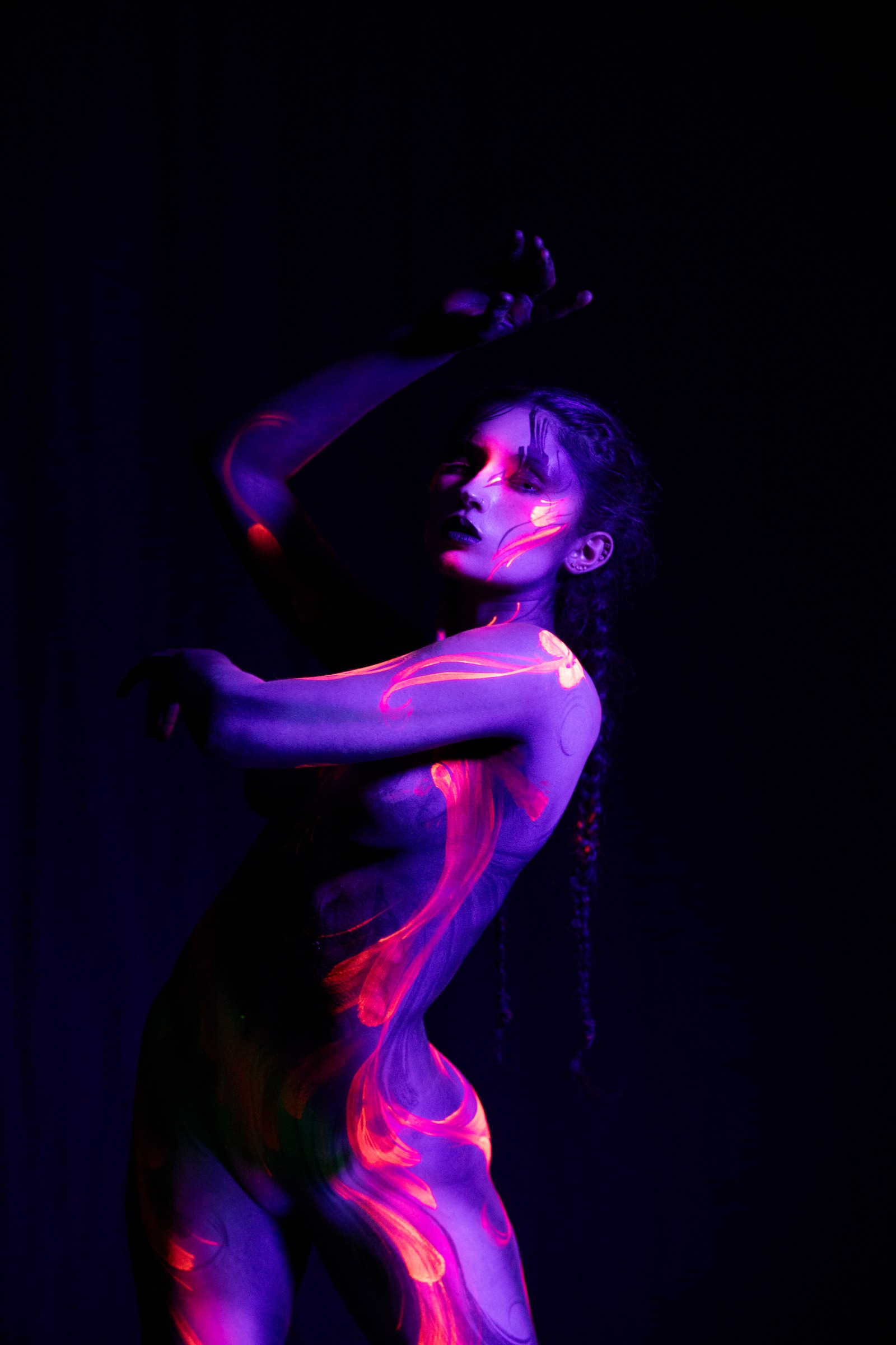 BabeyGianna stands facing the camera with her head tilted downward, shown in a close upper-body view. A purple UV base is overlaid with an orange and pink floral pattern centered on the chest and shoulder. Bodypaint by Claudia Kraemer. Photographed under blacklight by Elad Itzkin Photography in Gesundbrunnen, Berlin.