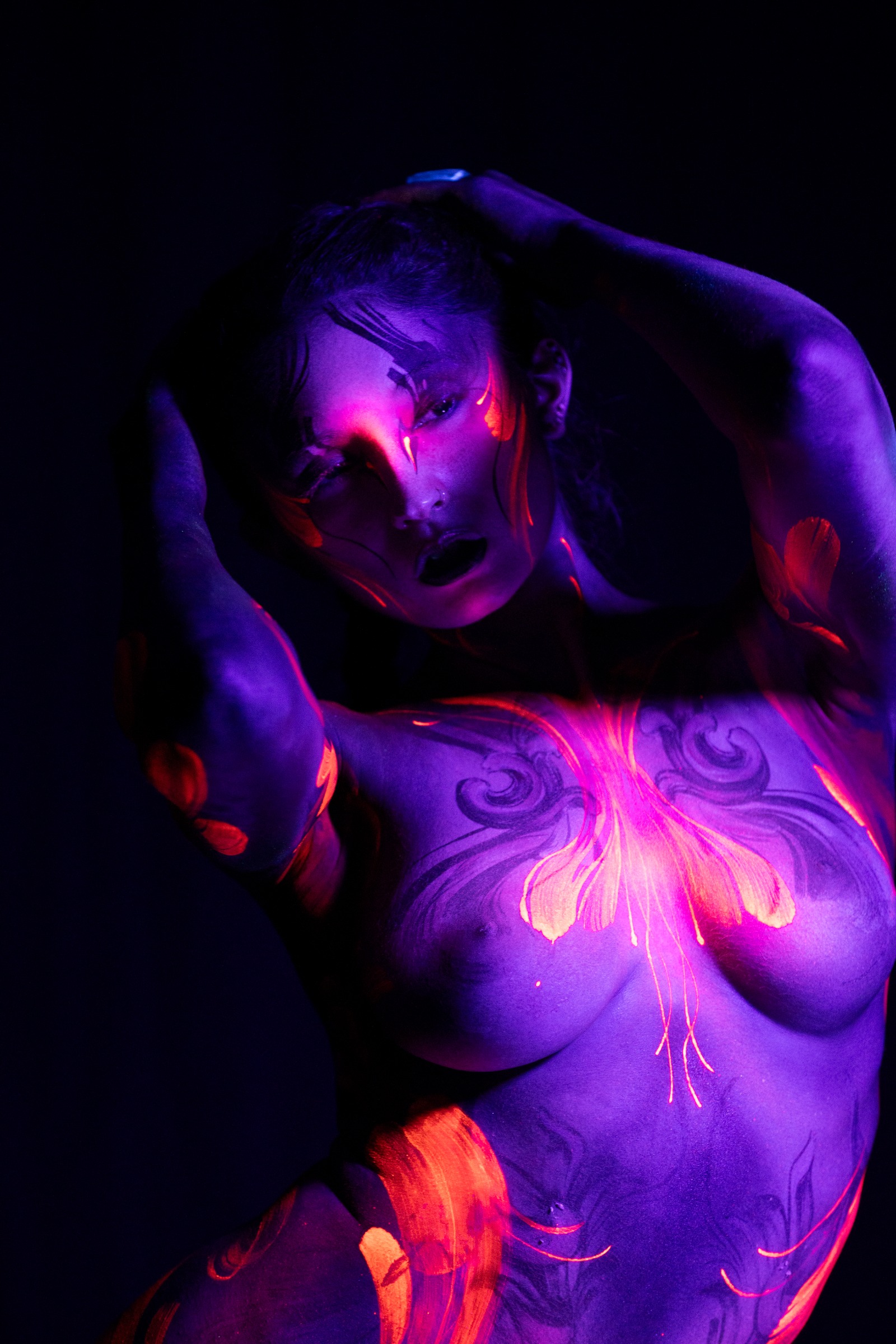 BabeyGianna stands facing the camera directly in a close upper-body view. Orange and red flame-like petal patterns wrap across a violet UV base on her chest and torso. Bodypaint by Claudia Kraemer. Photographed by Elad Itzkin Photography under UV light in his Berlin studio.