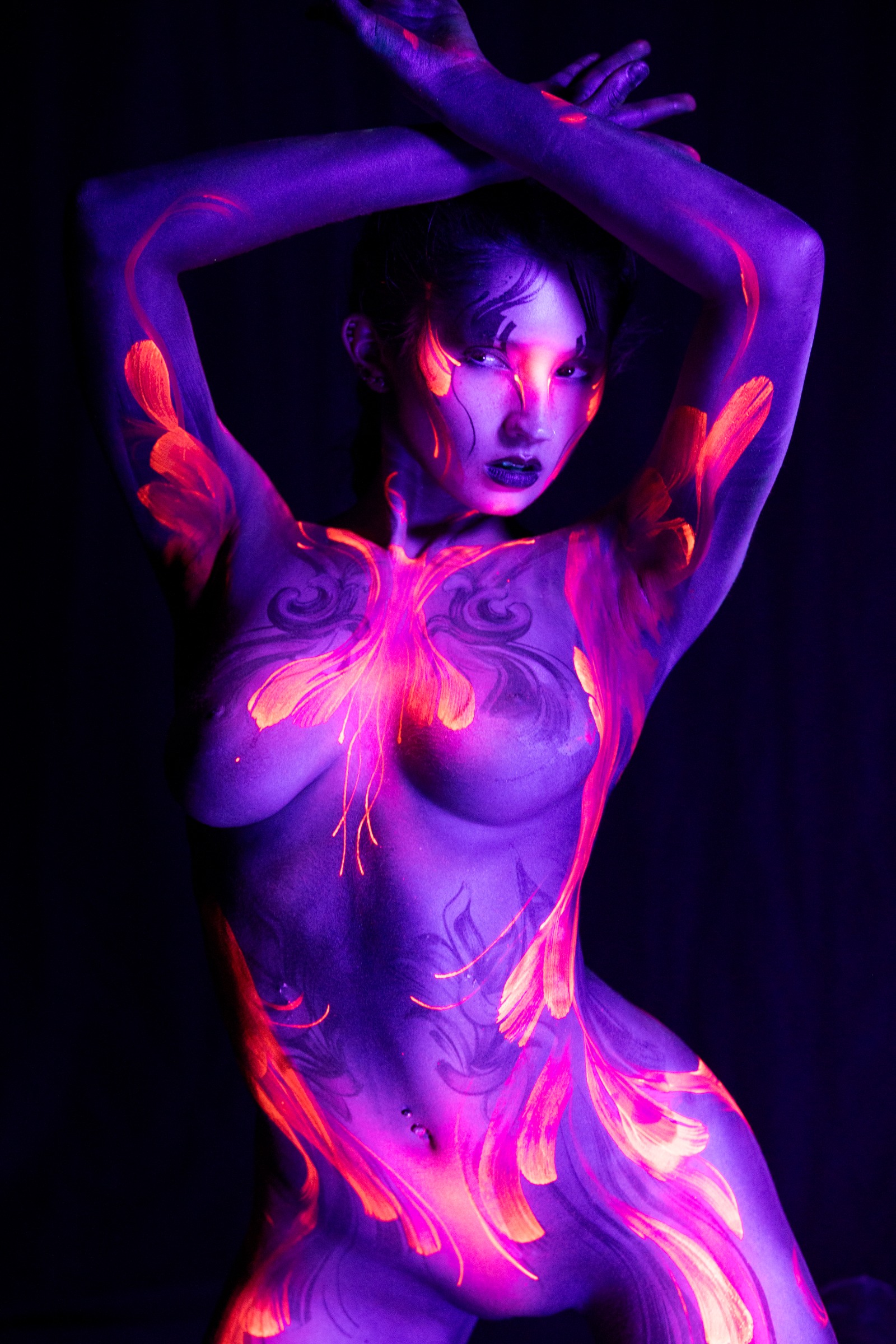 Standing and facing the camera directly, BabeyGianna is painted with a violet UV base and a bright orange and pink floral design centered on her chest and torso. Bodypaint by Claudia Kraemer. Photographed by Elad Itzkin Photography under UV light in his Berlin studio.