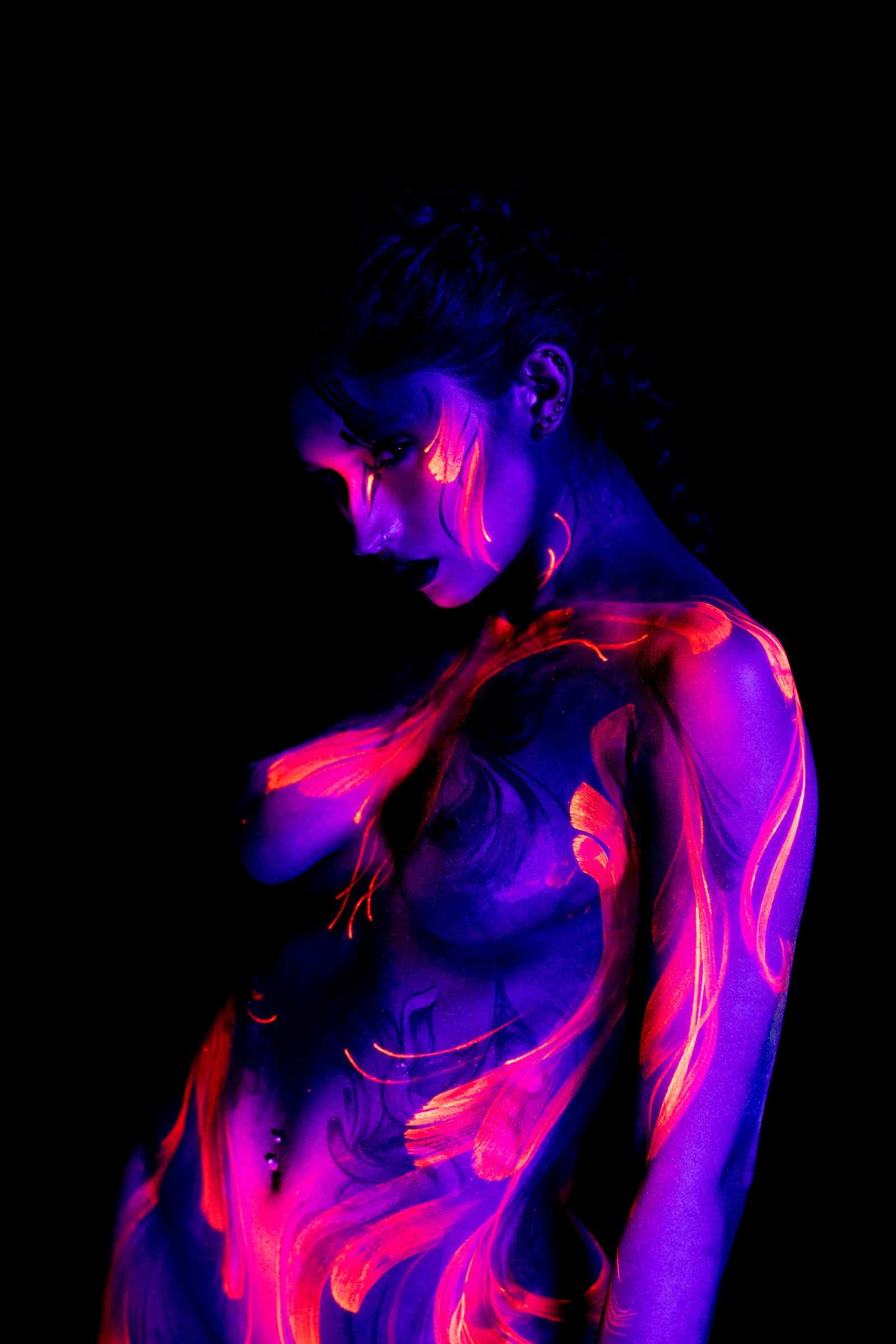 BabeyGianna is shown in a three-quarter side view with her head tilted downward. Pink and orange abstract linework flows across her chest and torso under UV light. Bodypaint by Claudia Kraemer. Photographed by Elad Itzkin Photography under UV blacklight in his Gesundbrunnen, Berlin studio.