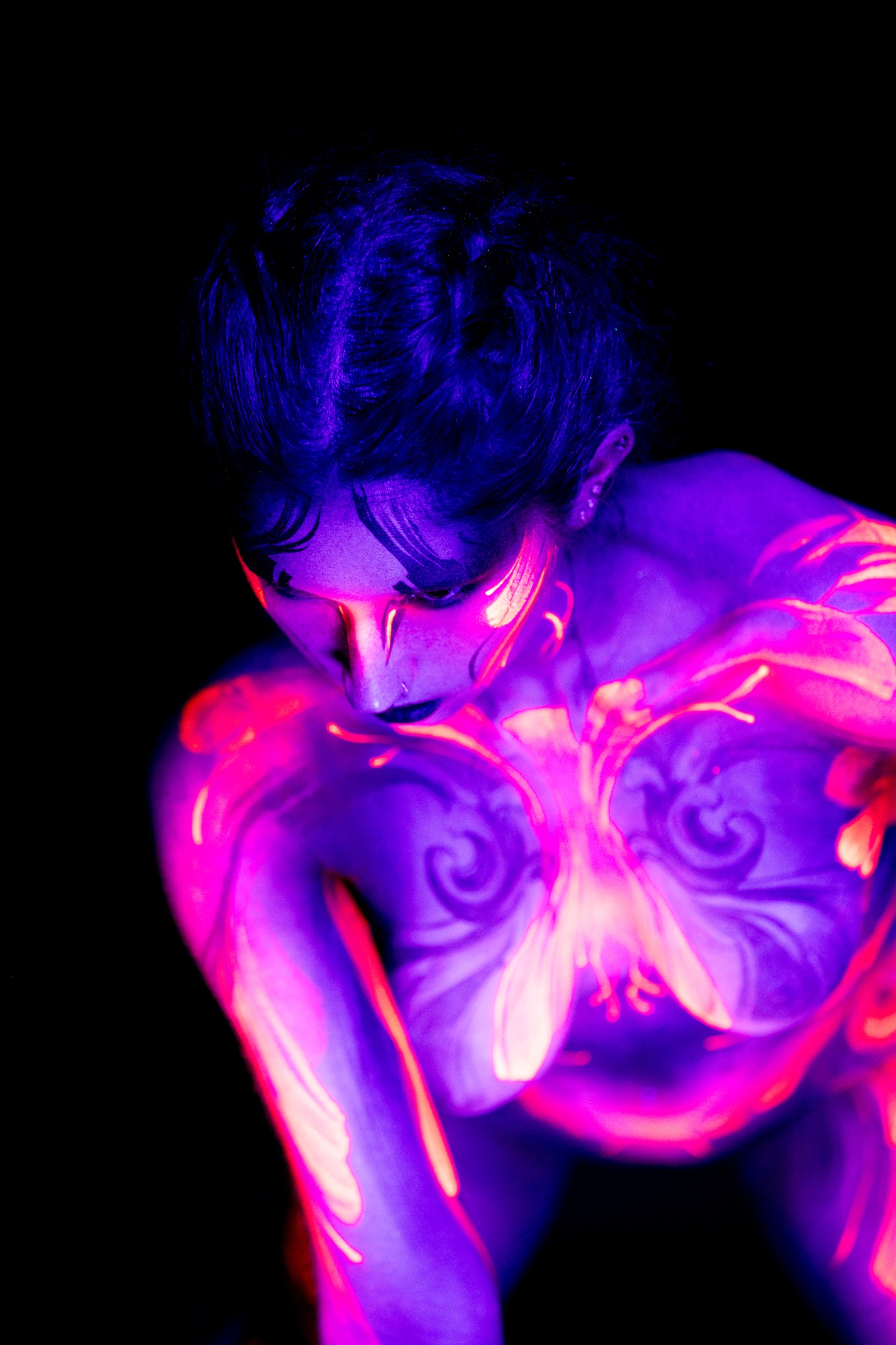 BabeyGianna is photographed bent slightly forward with her head lowered, shown in a close three-quarter view. A symmetrical pink and orange floral pattern with dark scrollwork covers her chest. Bodypaint by Claudia Kraemer. Photographed by Elad Itzkin Photography under UV blacklight in his studio in Gesundbrunnen, Berlin.