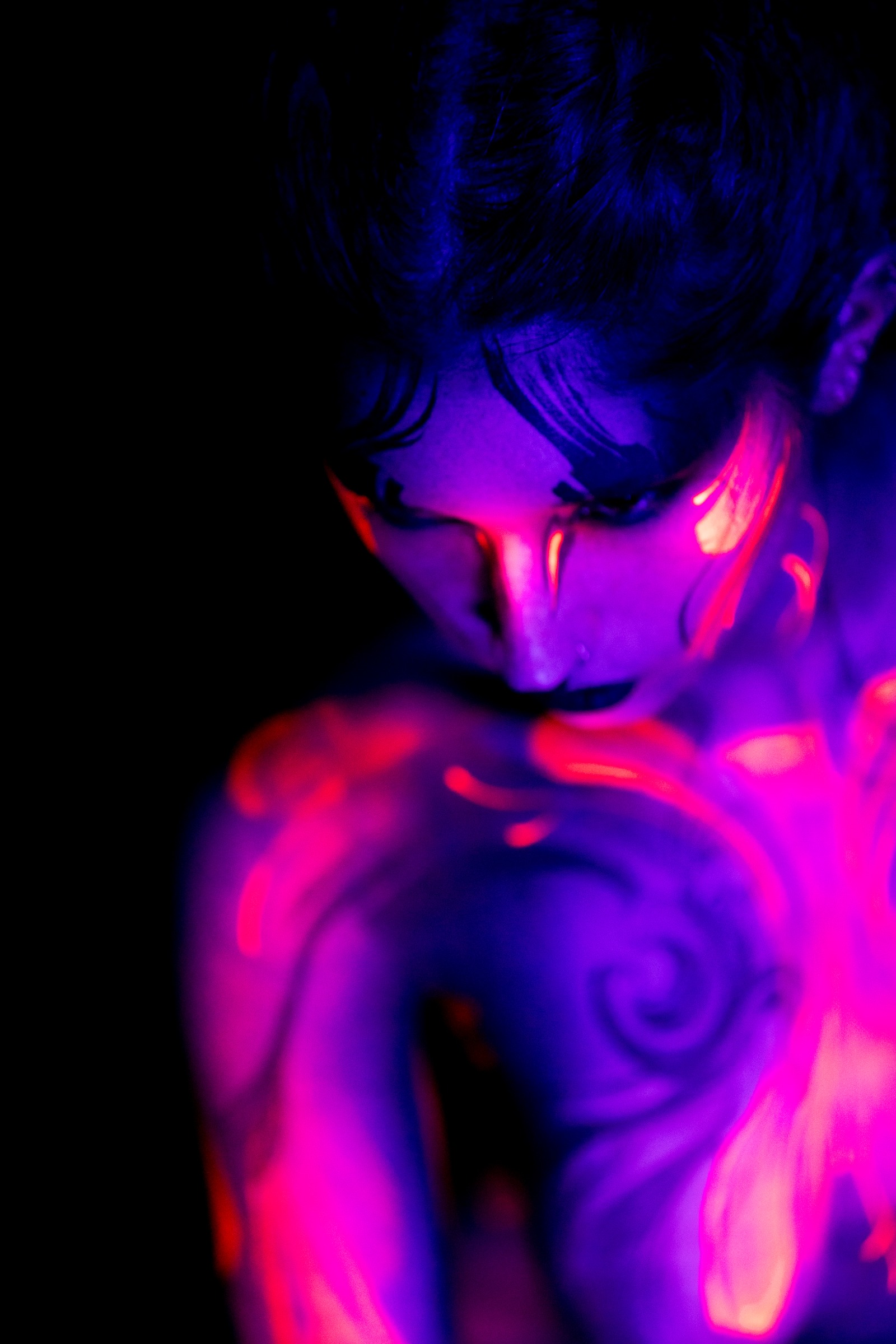 BabeyGianna stands with her head lowered in a close, softly focused view of her shoulder and collarbone. A red and orange swirl pattern glows across her skin under UV light. Bodypaint by Claudia Kraemer. Photographed by Elad Itzkin Photography in his Gesundbrunnen, Berlin studio.