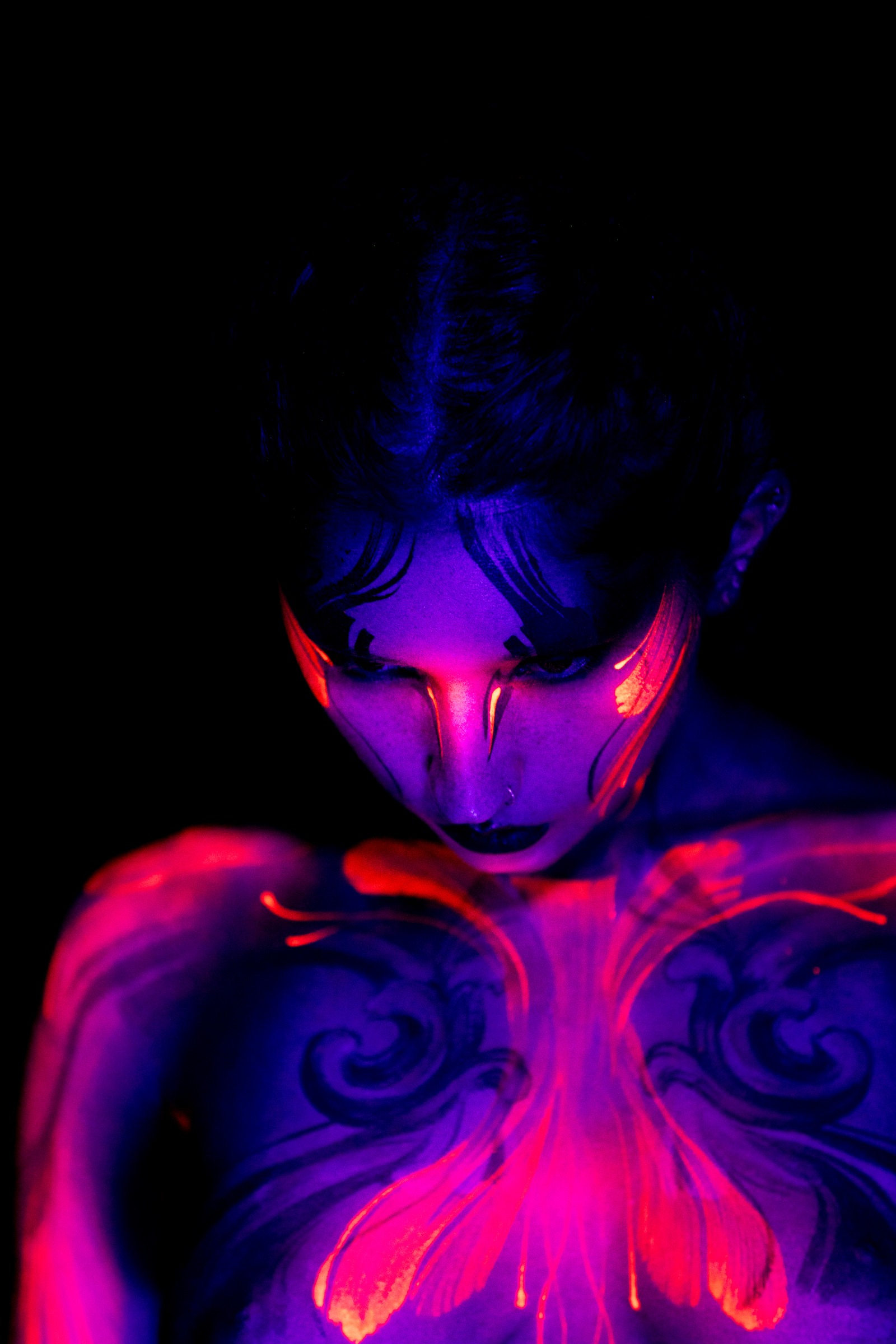 BabeyGianna stands facing forward with her head tilted down, framed from the chest up. A symmetrical pink and orange floral design bordered with dark scrollwork spans her chest. Bodypaint by Claudia Kraemer. Photographed by Elad Itzkin Photography under UV blacklight in his Gesundbrunnen, Berlin studio.