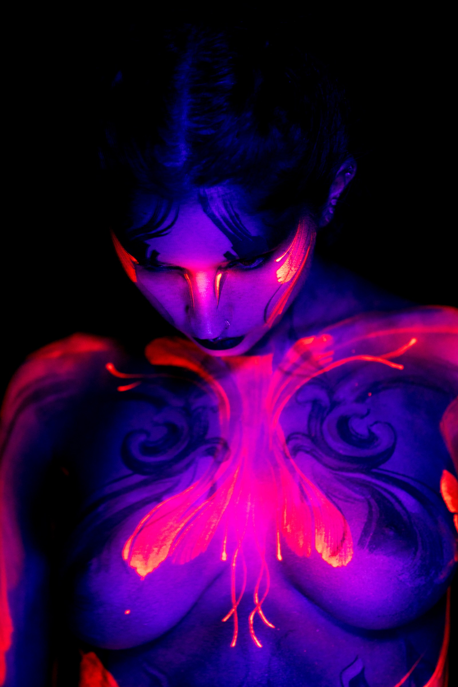 BabeyGianna stands facing the camera with her head lowered. A symmetrical floral design in pink and orange, outlined with dark scrollwork, covers her chest and collarbone. Bodypaint by Claudia Kraemer. Photographed under UV blacklight by Elad Itzkin Photography in his Gesundbrunnen, Berlin studio.