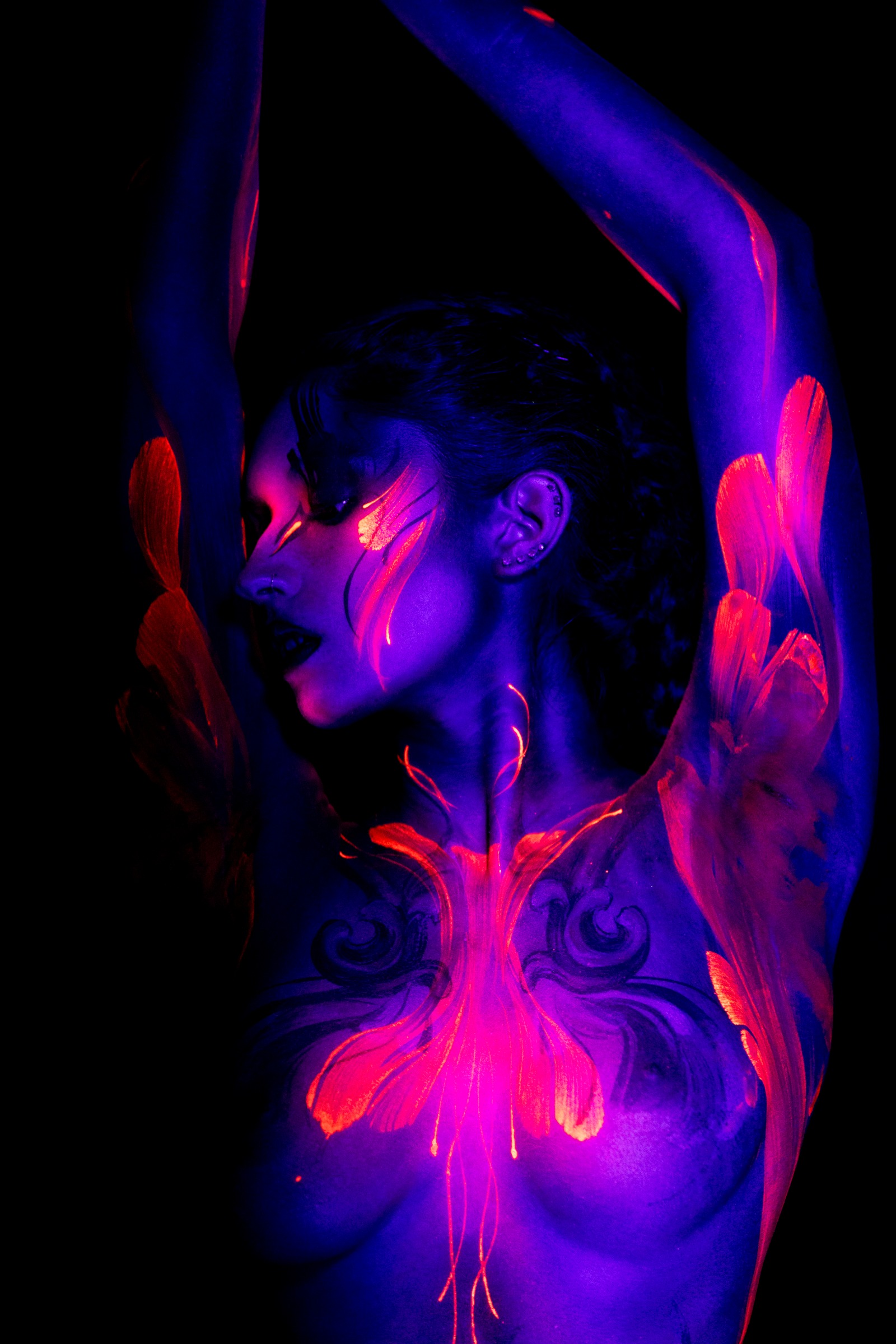 BabeyGianna is shown in profile with her head turned to the side. Orange feather-like patterns extend along her torso beside a pink and orange floral design centered on her chest. Bodypaint by Claudia Kraemer. Photographed by Elad Itzkin Photography under UV blacklight in his Gesundbrunnen, Berlin studio.