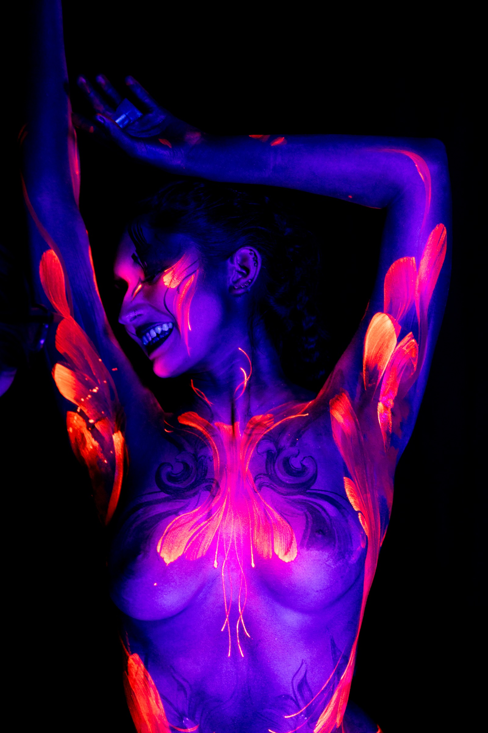 BabeyGianna stands with her head tilted back, captured in a three-quarter view. Orange feather-like brushstrokes frame a pink and orange floral pattern across her chest under UV light. Bodypaint by Claudia Kraemer. Photographed by Elad Itzkin Photography under UV blacklight in his Gesundbrunnen, Berlin studio.