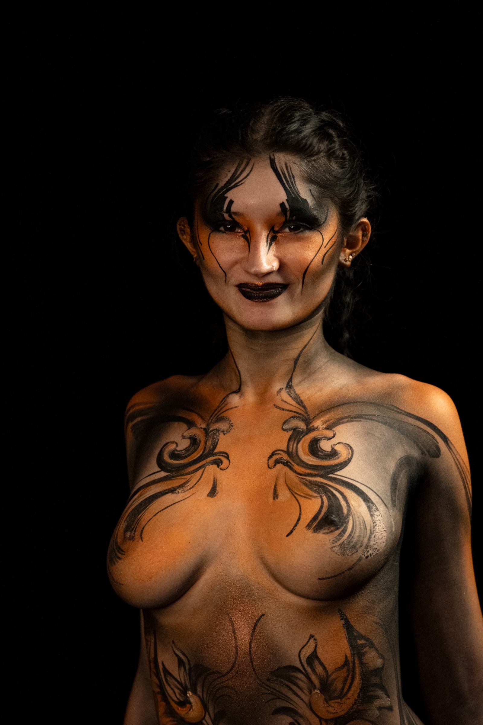 BabeyGianna stands facing the camera in a close chest-up view. Her torso is decorated with an orange and black abstract floral linework pattern. Bodypaint by Claudia Kraemer. Photographed by Elad Itzkin Photography under UV/blacklight in his studio in Gesundbrunnen, Berlin.