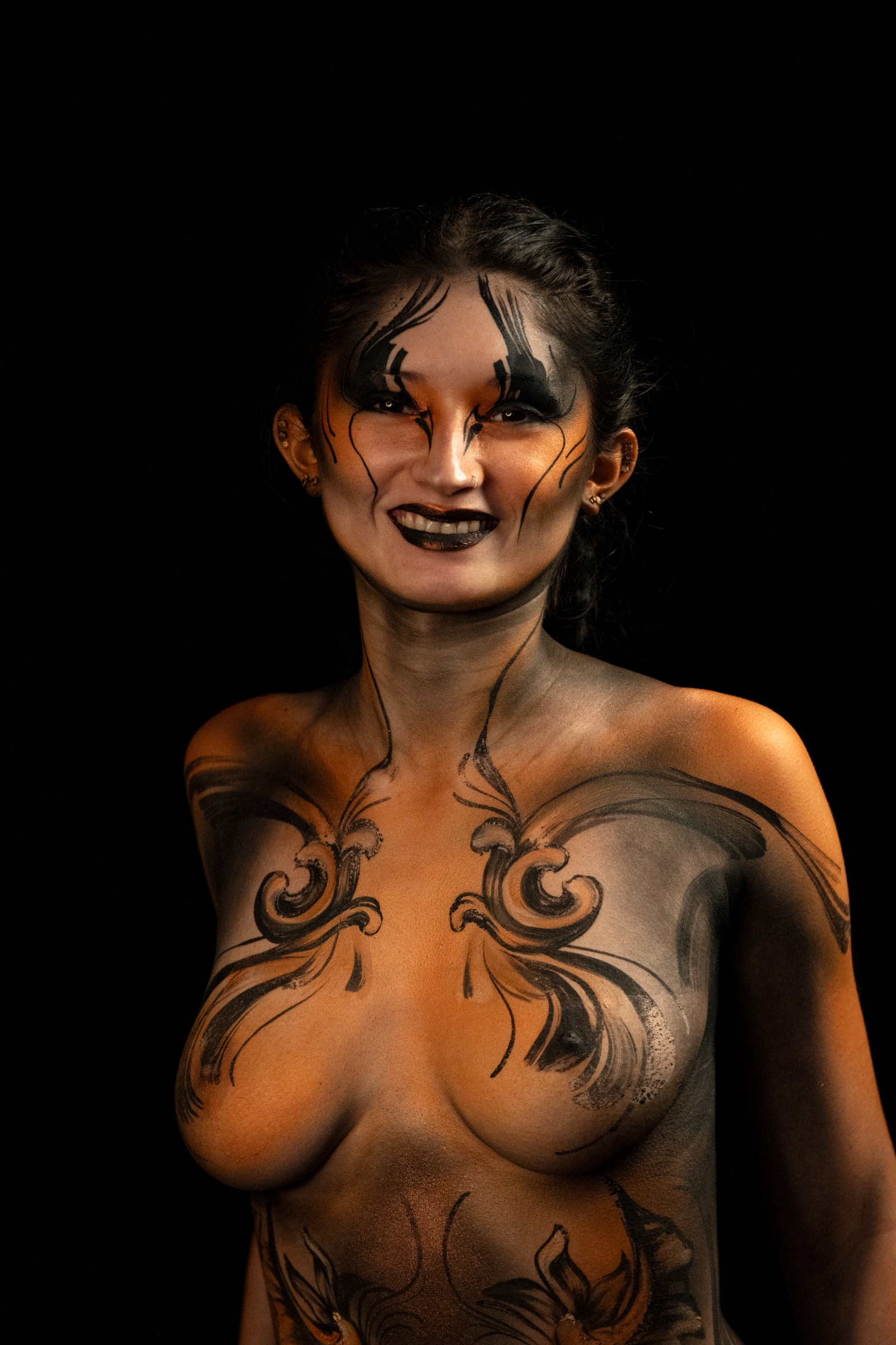 BabeyGianna stands in a three-quarter pose, framed from the chest up. The bodypaint spreads across the chest and shoulders in an orange and black abstract linework pattern. Bodypaint by Claudia Kraemer. Photographed by Elad Itzkin Photography under UV/blacklight in his studio in Gesundbrunnen, Berlin.