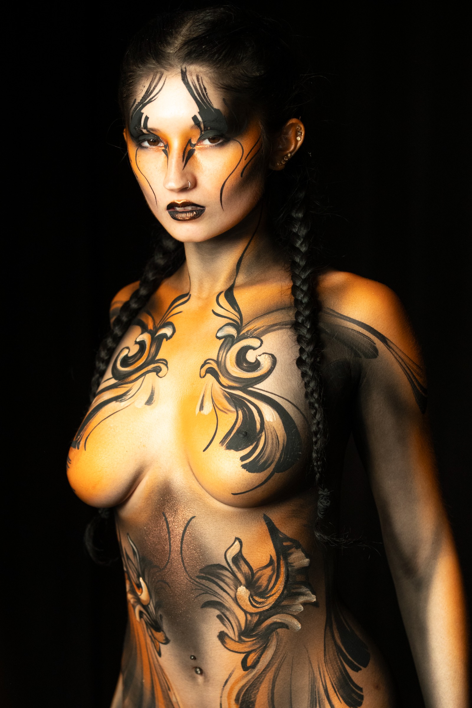BabeyGianna stands facing the camera, shown from the chest up with her hair in braids. Her body is painted in an orange and black abstract linework design with a swirling floral pattern. Bodypaint by Claudia Kraemer. Photographed by Elad Itzkin Photography under UV/blacklight in his studio in Gesundbrunnen, Berlin.