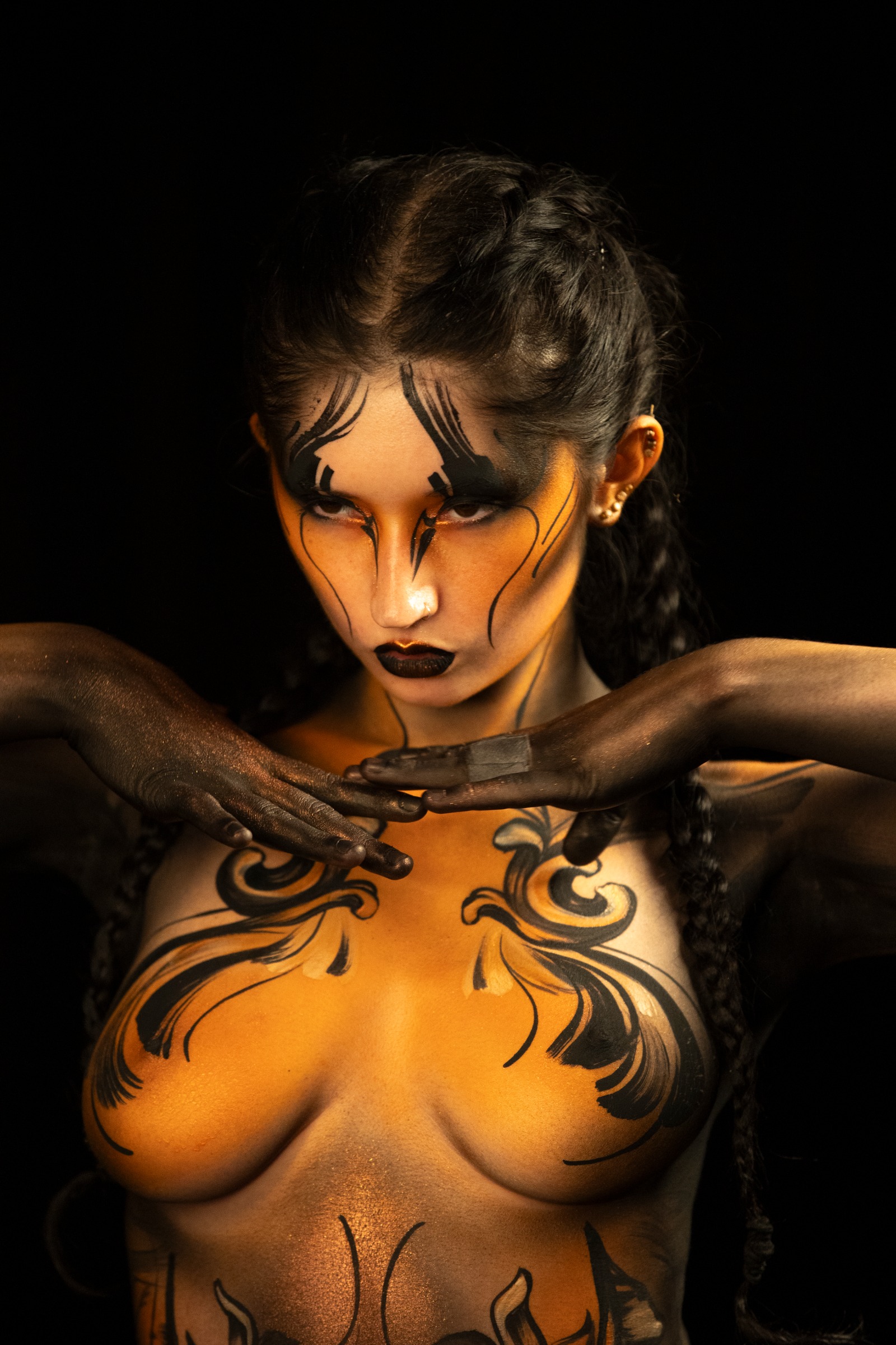 BabeyGianna stands facing the camera with her head lowered slightly. Orange and black swirling linework covers her torso in a symmetrical scrollwork design. Bodypaint by Claudia Kraemer. Photographed by Elad Itzkin Photography under UV blacklight in his studio in Gesundbrunnen, Berlin.