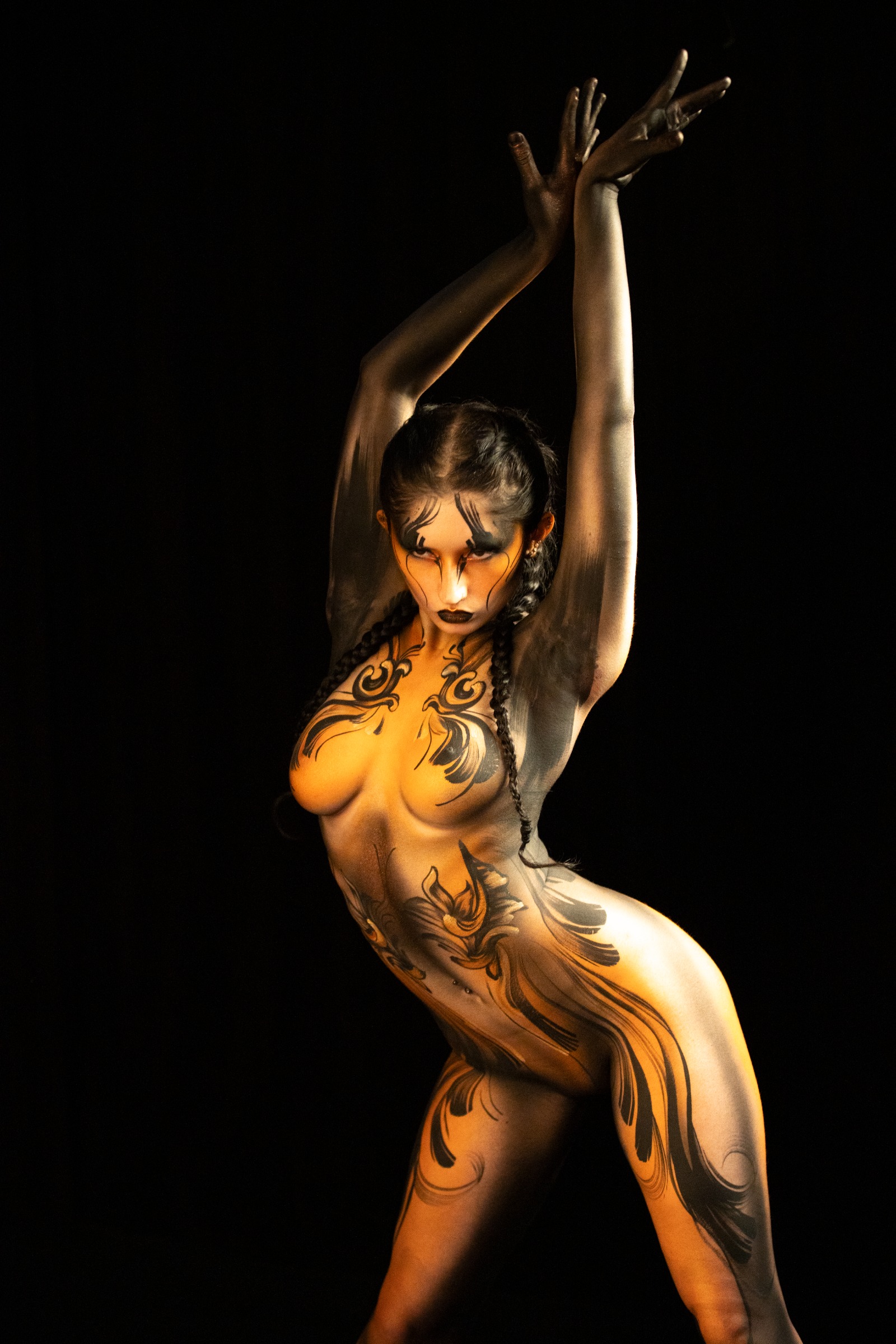 BabeyGianna stands in a three-quarter pose with her body turned toward the side. Orange and black scrollwork linework covers her torso in a symmetrical floral pattern. Bodypaint by Claudia Kraemer. Photographed by Elad Itzkin Photography under UV blacklight in his studio in Gesundbrunnen, Berlin.