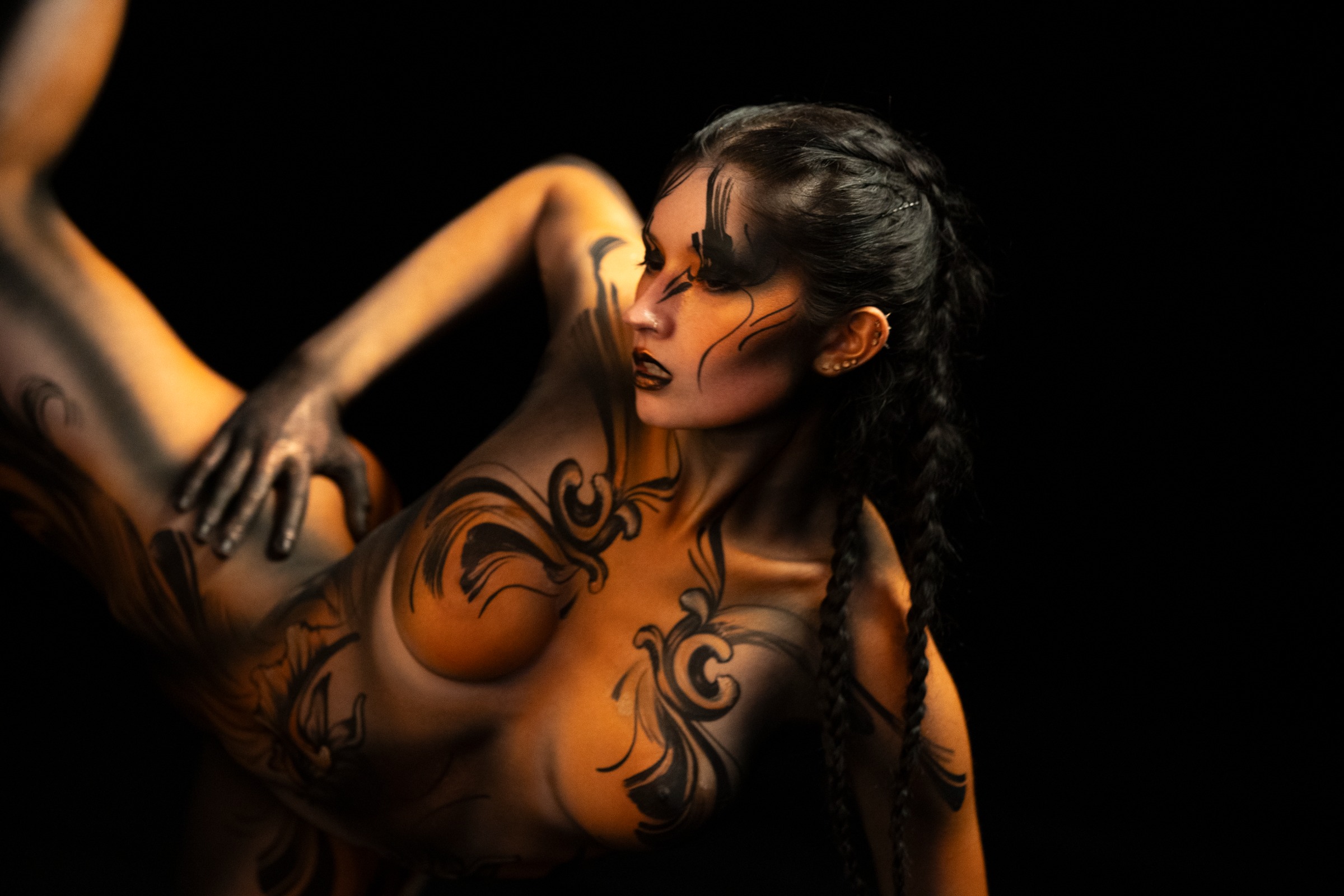 BabeyGianna is photographed leaning back with her torso arched in a twisting three-quarter pose. The design uses orange and black tones in a tribal scrollwork pattern across the chest and shoulder. Bodypaint by Claudia Kraemer. Photographed by Elad Itzkin Photography under UV light in his Gesundbrunnen, Berlin studio.