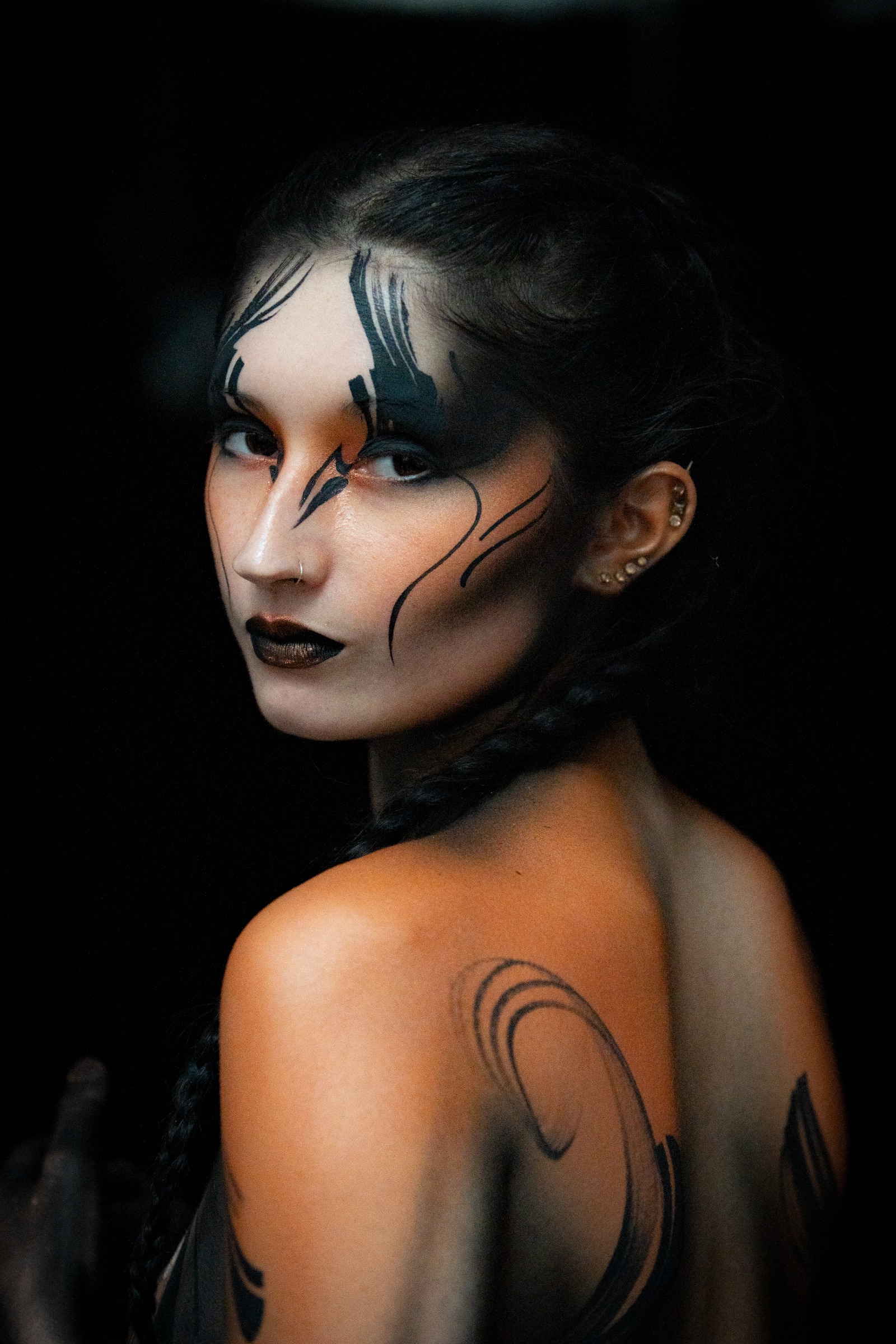 BabeyGianna is photographed with her back turned to the camera, looking back over her shoulder. Black swirl linework in orange and black tones decorates her shoulder and upper back, with matching dark linework on her face. Bodypaint by Claudia Kraemer. Photographed by Elad Itzkin Photography under UV light in his Gesundbrunnen, Berlin studio.
