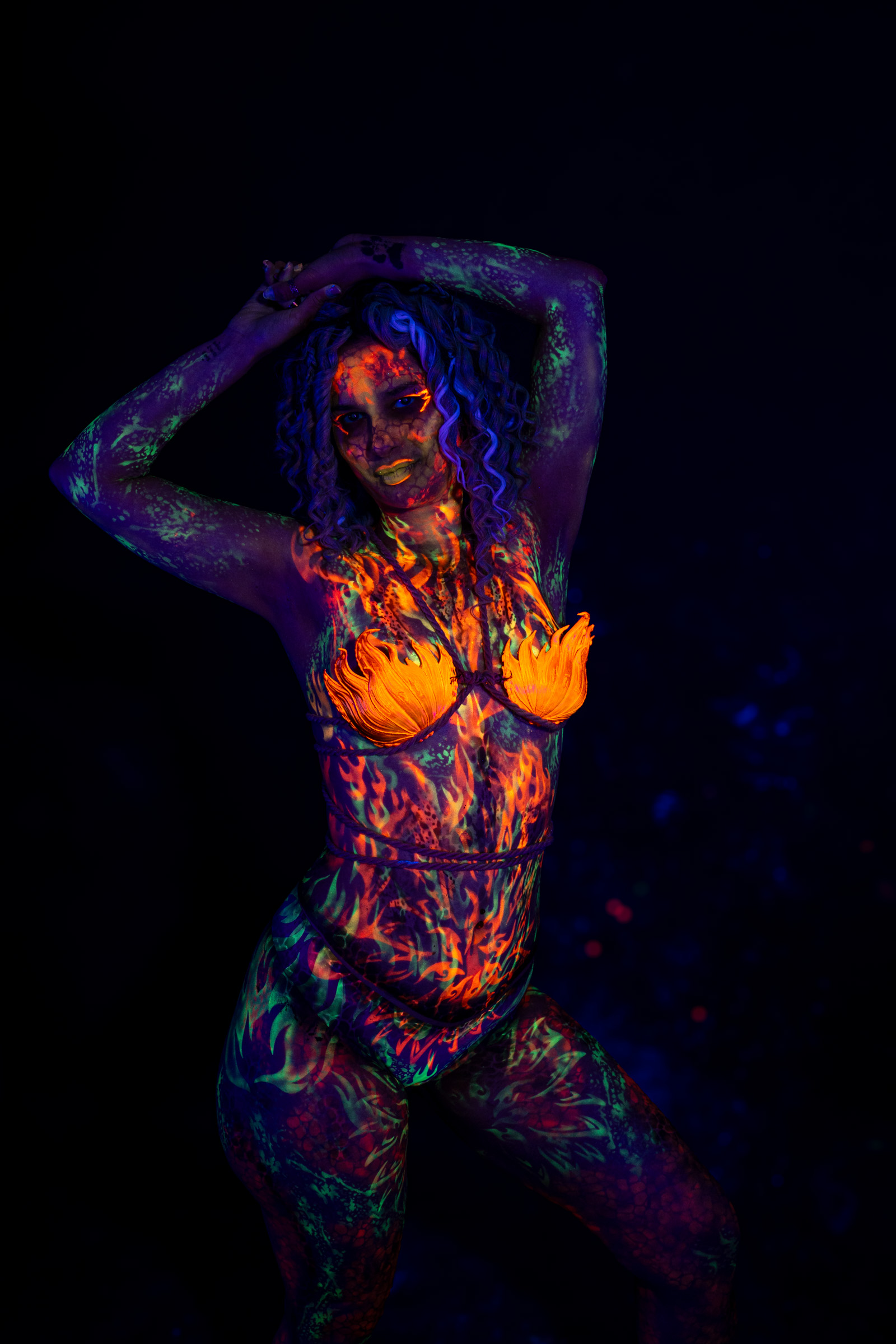 Bella stands facing the camera at a slight angle. Her body is painted in an orange, red, green, and blue flame-like tribal print pattern covering her torso and legs. Bodypaint by Elad Itzkin. Photographed by Elad Itzkin Photography under UV/blacklight in his studio in Gesundbrunnen, Berlin.