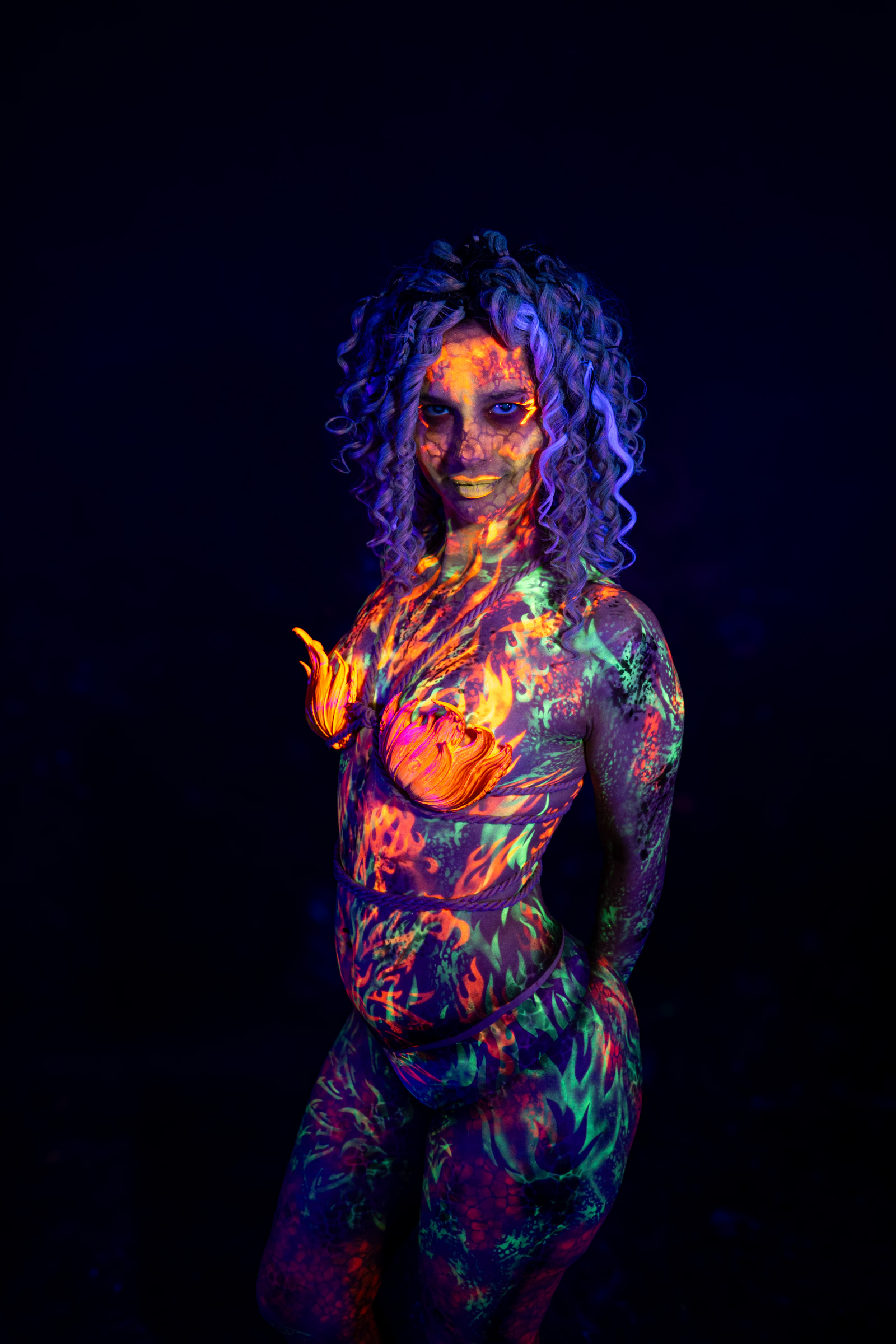 Bella stands facing the camera at a slight angle. Her body is painted with an orange abstract pattern across the torso and green and purple mottled paint on the limbs. Bodypaint by Elad Itzkin. Photographed by Elad Itzkin Photography under UV/blacklight in his studio in Gesundbrunnen, Berlin.