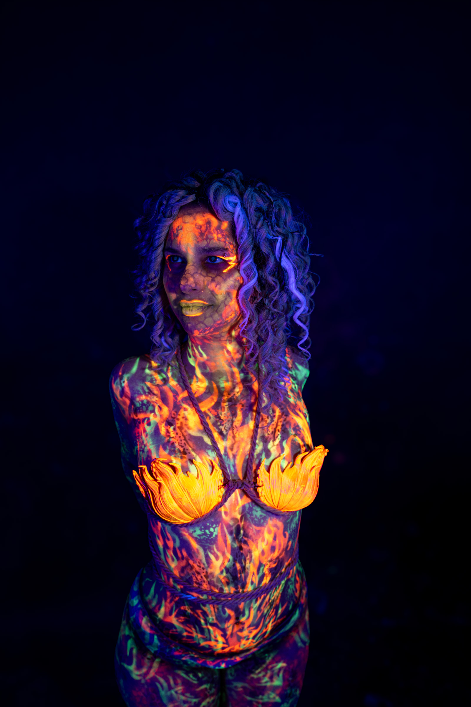 Bella stands facing forward, turned slightly to one side. An orange flame petal pattern covers her chest, and an abstract orange and green design continues across her torso and legs. Bodypaint by Elad Itzkin. Photographed by Elad Itzkin Photography under UV/blacklight in his studio in Gesundbrunnen, Berlin.