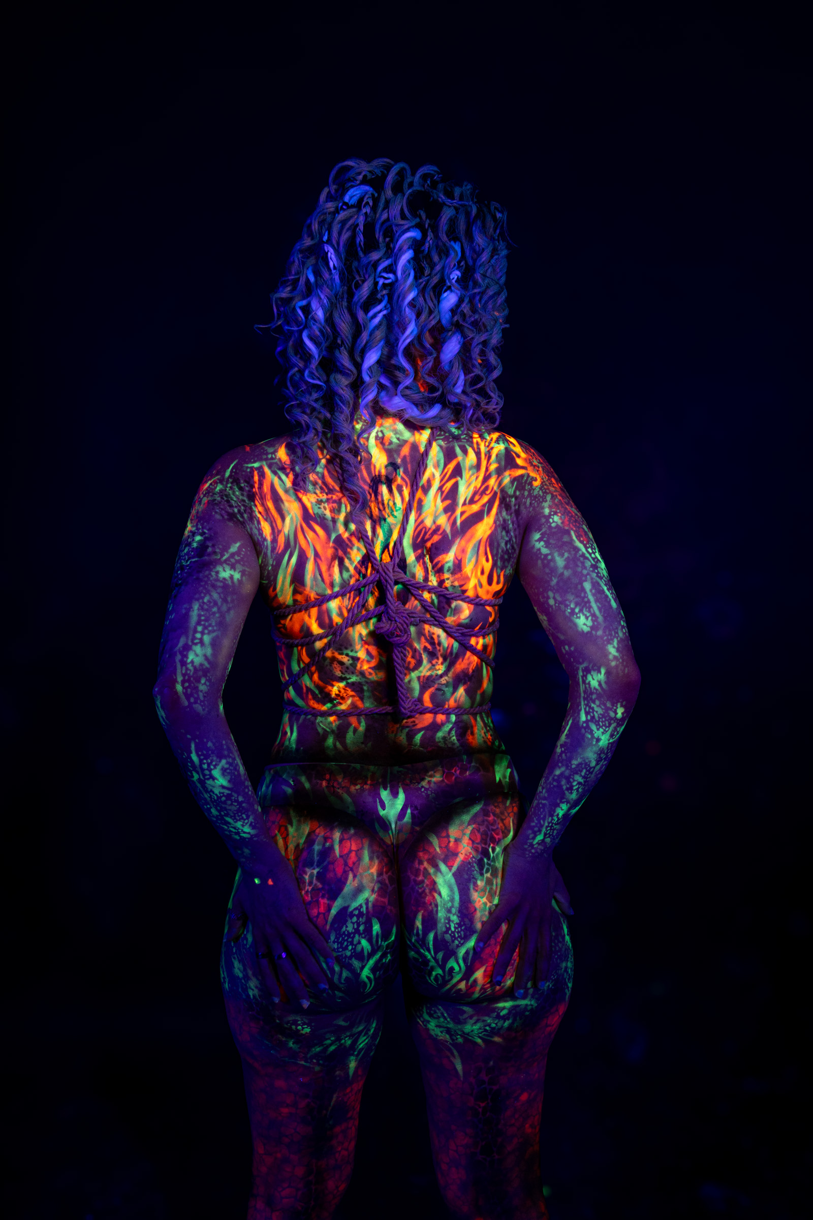 Bella stands with her back to the camera, her full body visible from behind. Her back and legs carry an abstract orange, red, and green pattern. Bodypaint by Elad Itzkin. Photographed by Elad Itzkin Photography under UV/blacklight in his studio in Gesundbrunnen, Berlin.