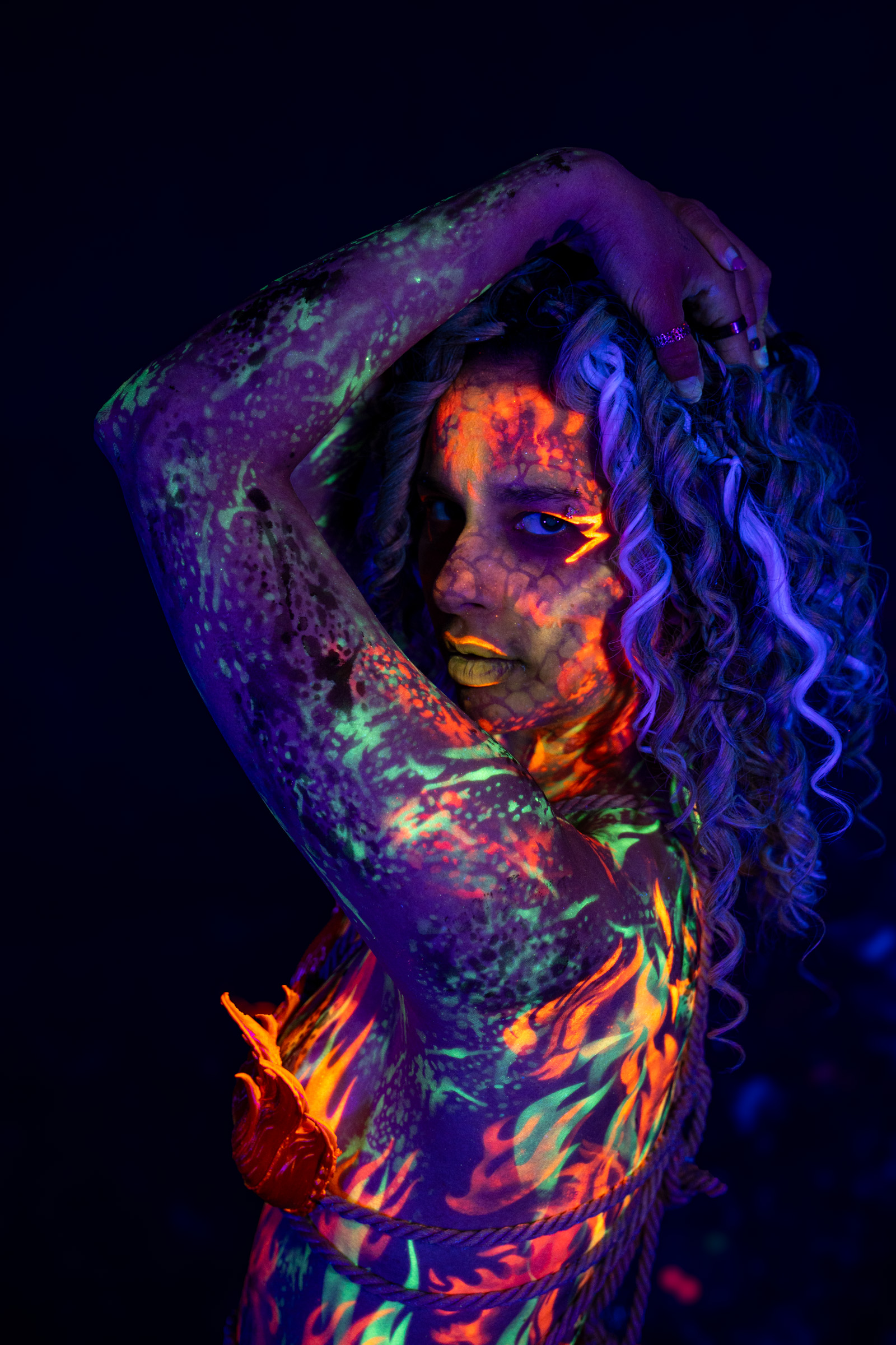 Model Bella is shown from the chest up in a close three-quarter pose, her head tilted to the side. An orange, red, and green mottled pattern covers her face and shoulder. Bodypaint by Elad Itzkin. Photographed by Elad Itzkin Photography under UV/blacklight in his studio in Gesundbrunnen, Berlin.