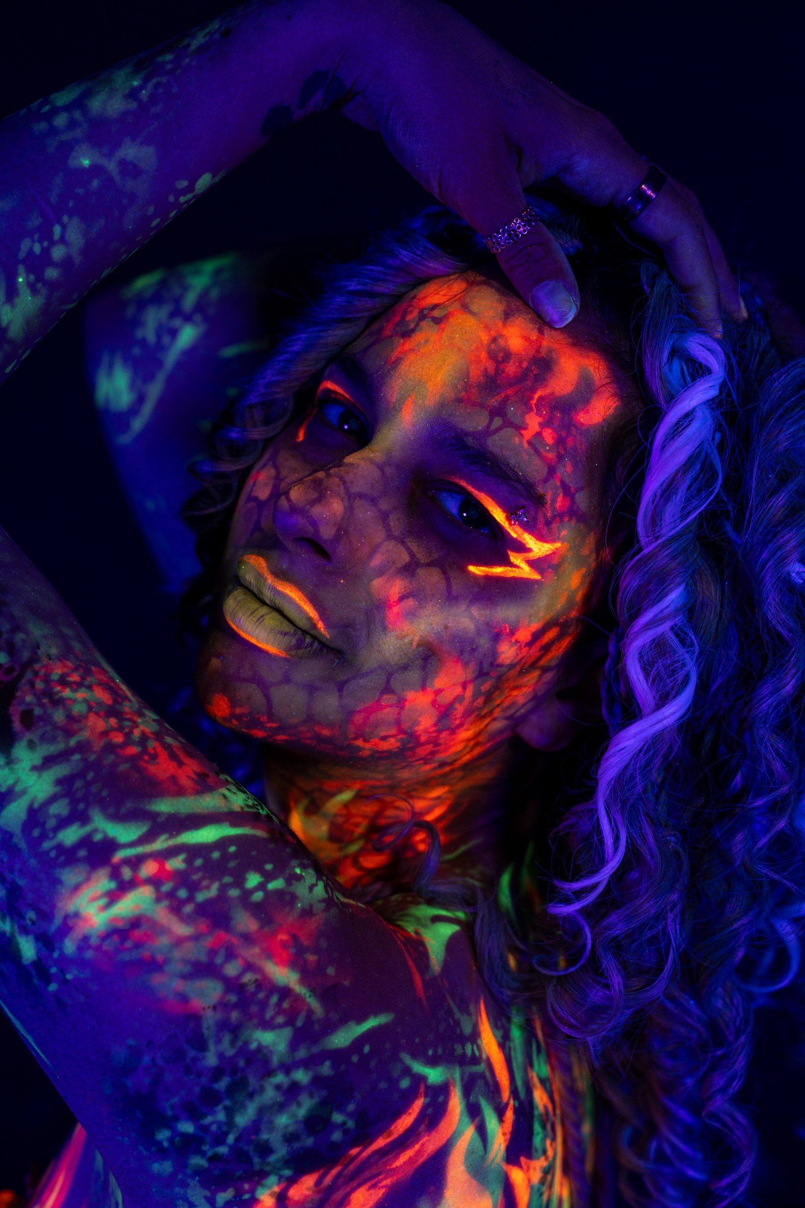 Bella is shown in a close three-quarter portrait with her head tilted back. Her face is painted with an orange and red netted pattern, and her shoulder carries a green mottled design. Bodypaint by Elad Itzkin. Photographed by Elad Itzkin Photography under UV/blacklight in his studio in Gesundbrunnen, Berlin.