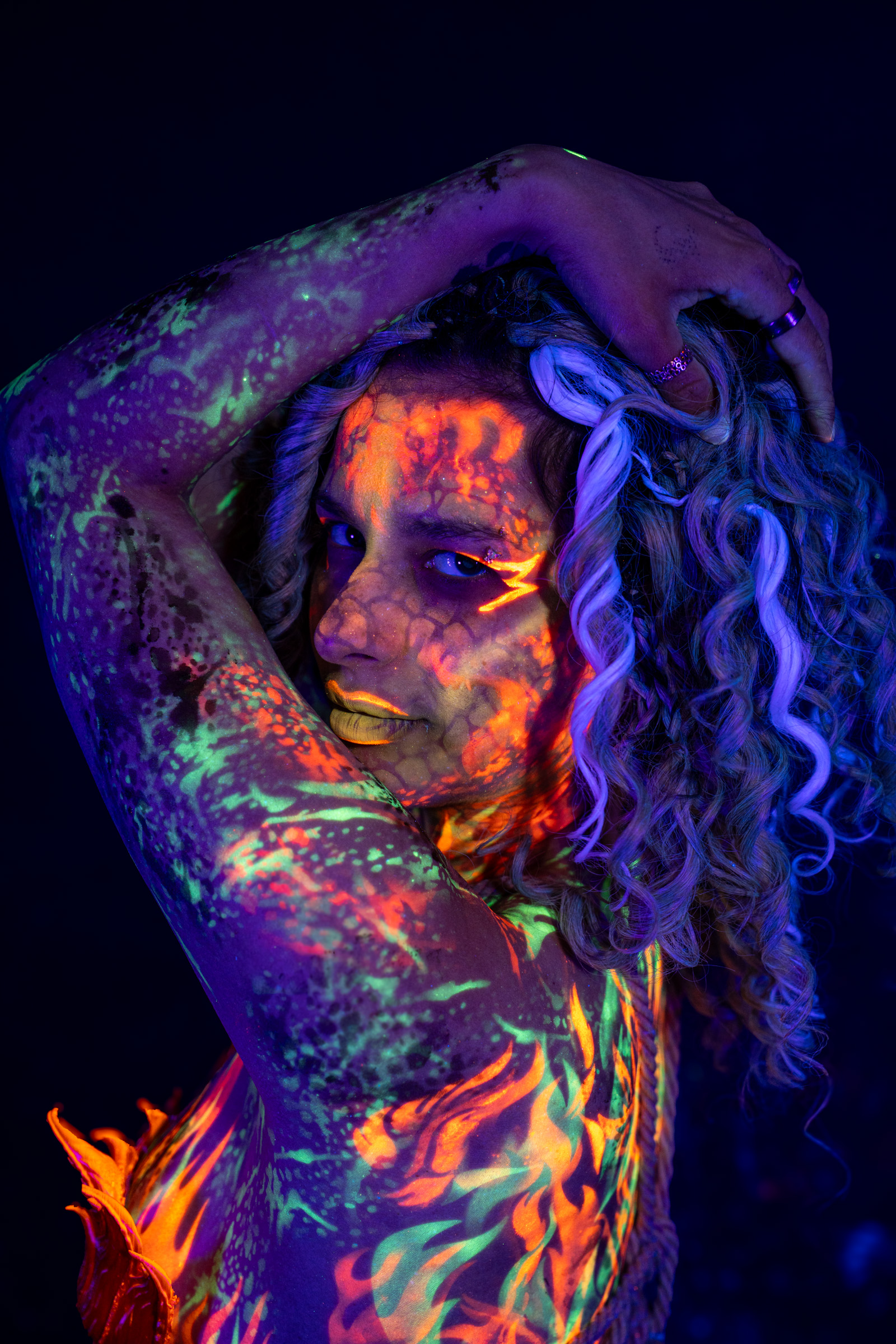 Model Bella is shown in a close three-quarter portrait with her head tilted back. An orange and red netted pattern covers her face, and green and purple mottled paint spreads across her shoulder. Bodypaint by Elad Itzkin. Photographed by Elad Itzkin Photography under UV/blacklight in his studio in Gesundbrunnen, Berlin.
