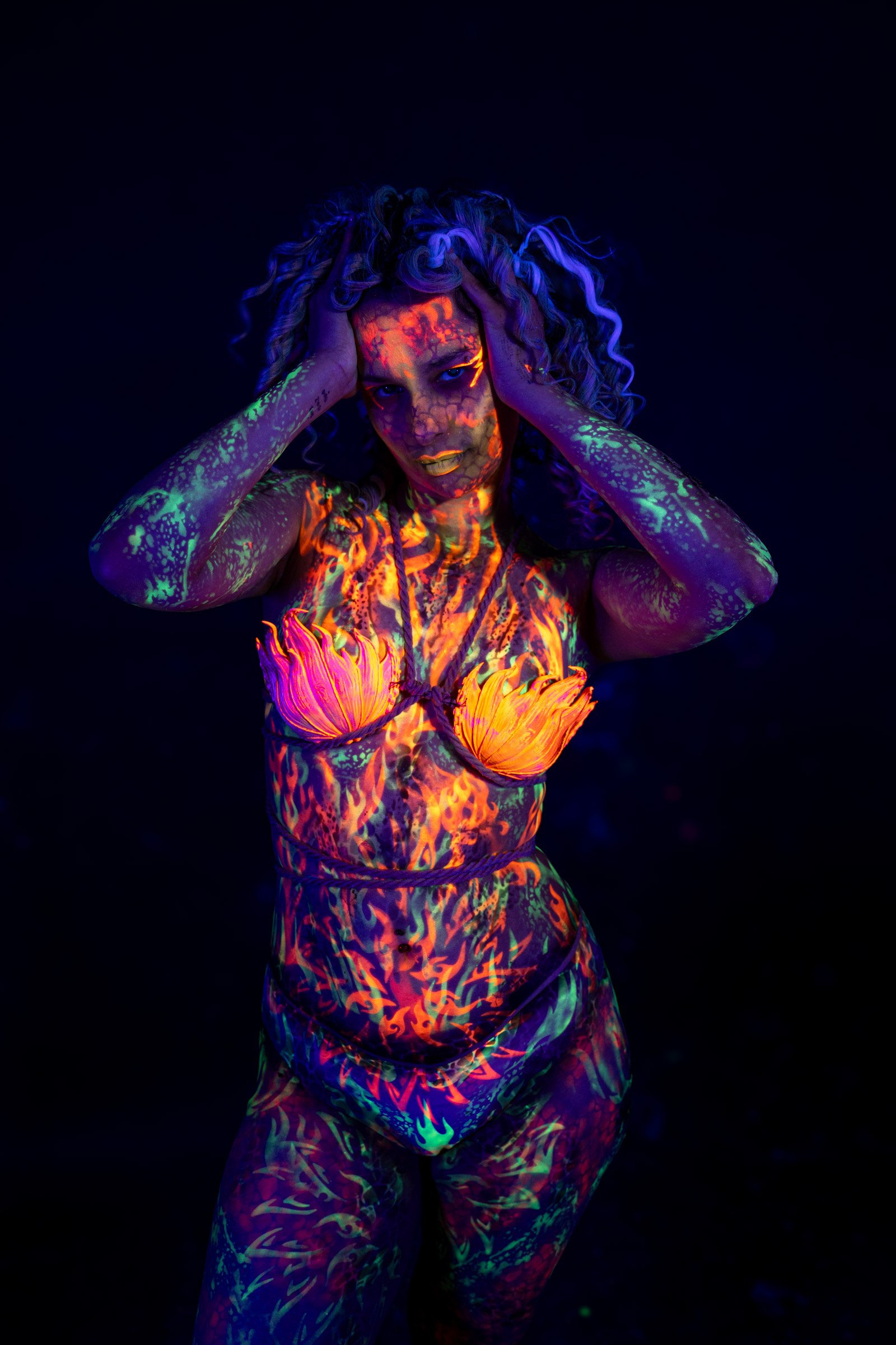 Model Bella stands facing the camera with her head tilted to one side. Her face carries a mottled orange and green pattern, and her chest is painted with an orange flame petal design. Bodypaint by Elad Itzkin. Photographed by Elad Itzkin Photography under UV/blacklight in his studio in Gesundbrunnen, Berlin.