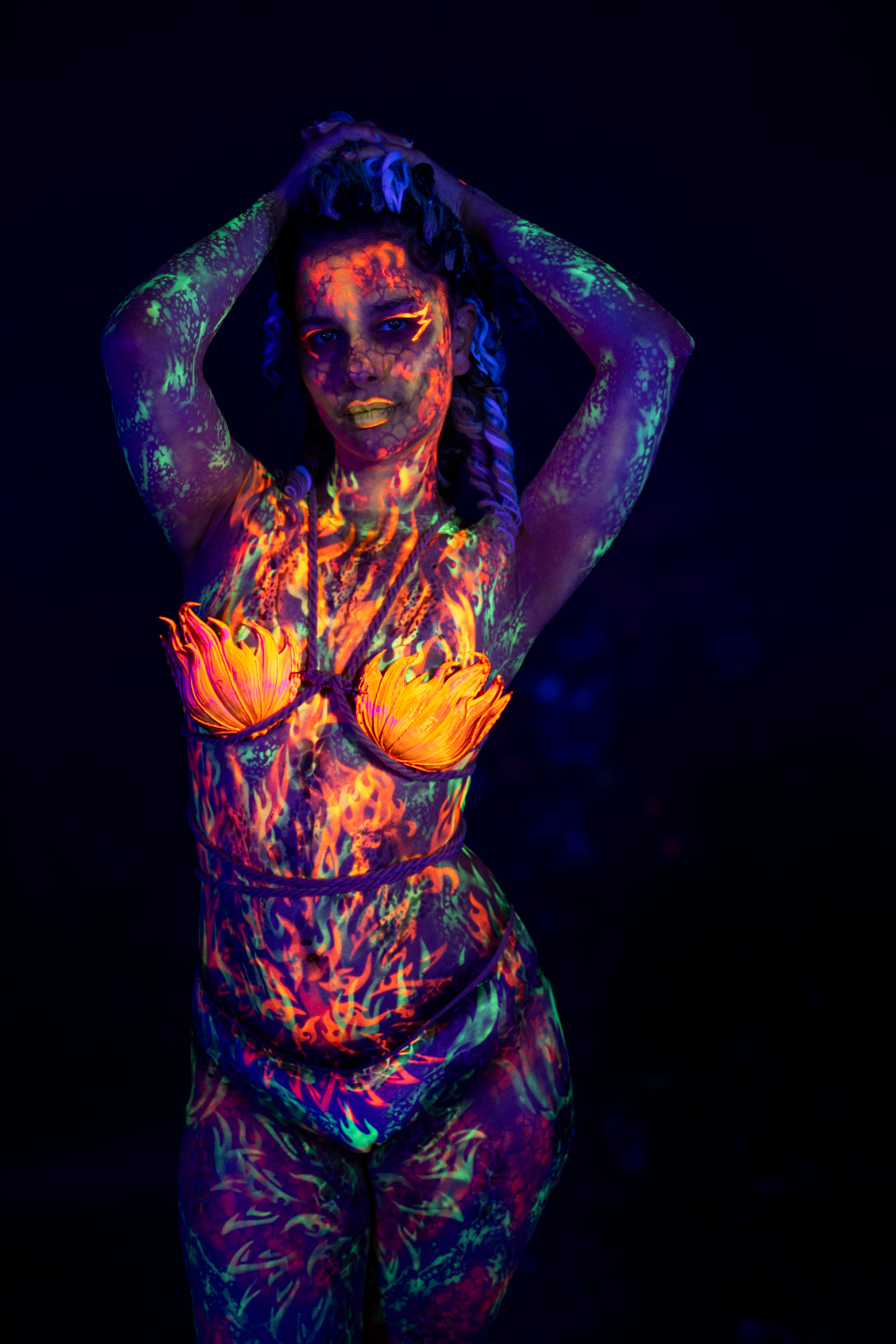 Model Bella stands facing the camera in a front-on pose. Her face is painted with a mottled orange and green pattern, and an orange flame petal design covers her chest. Bodypaint by Elad Itzkin. Photographed by Elad Itzkin Photography under UV/blacklight in his studio in Gesundbrunnen, Berlin.