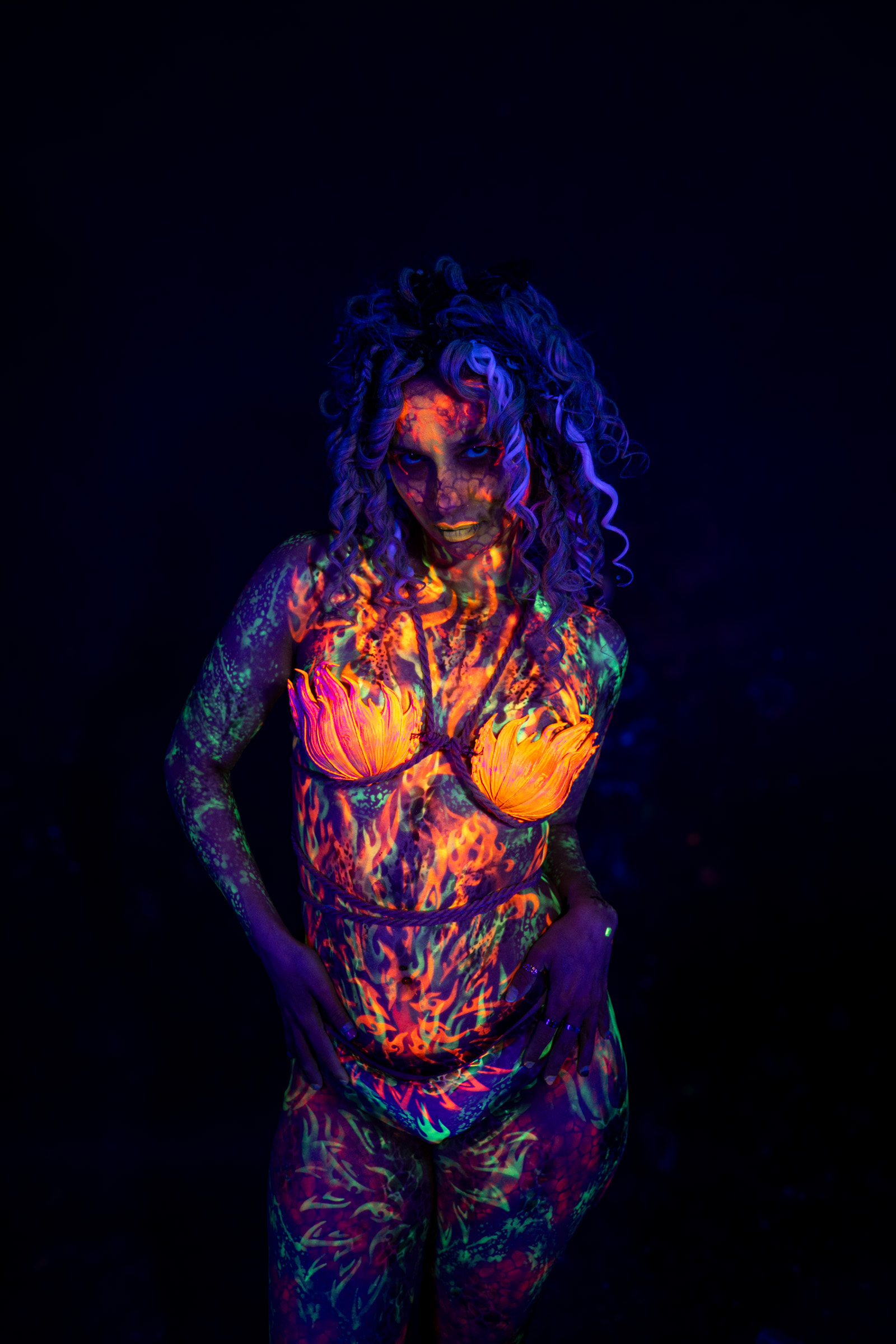 Bella stands facing the camera in a three-quarter view. Her body is painted in an orange and red flame-like pattern with green accents on the arms and an orange flower-shaped design over the chest. Bodypaint by Elad Itzkin. Photographed by Elad Itzkin Photography under UV light in his studio in Gesundbrunnen, Berlin.