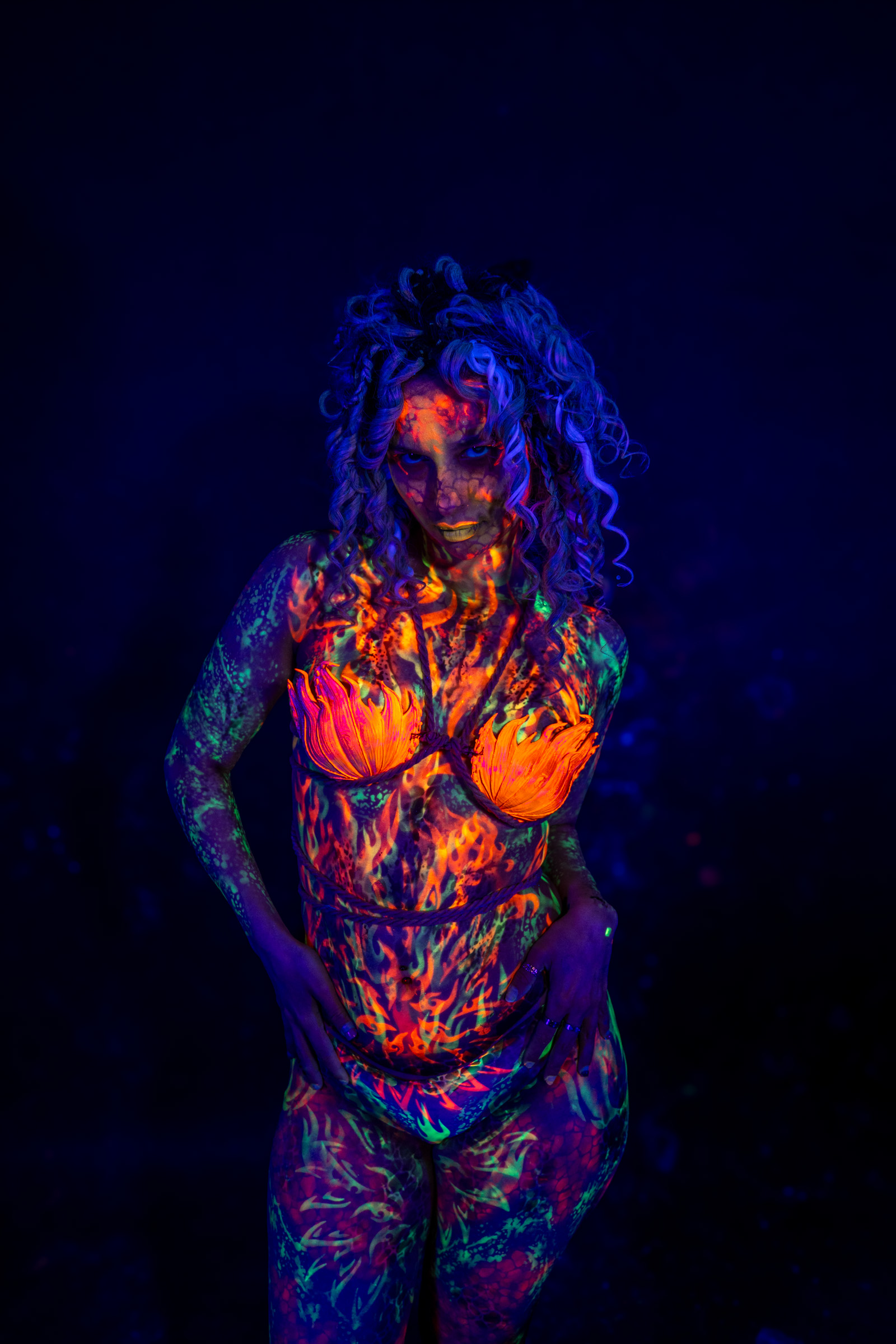 Bella stands in a three-quarter pose facing the camera, curly hair loose around her face. The bodypaint is a flame-inspired design in orange and red tones with green linework on the arms. Bodypaint by Elad Itzkin. Photographed by Elad Itzkin Photography under blacklight in his studio in Gesundbrunnen, Berlin.