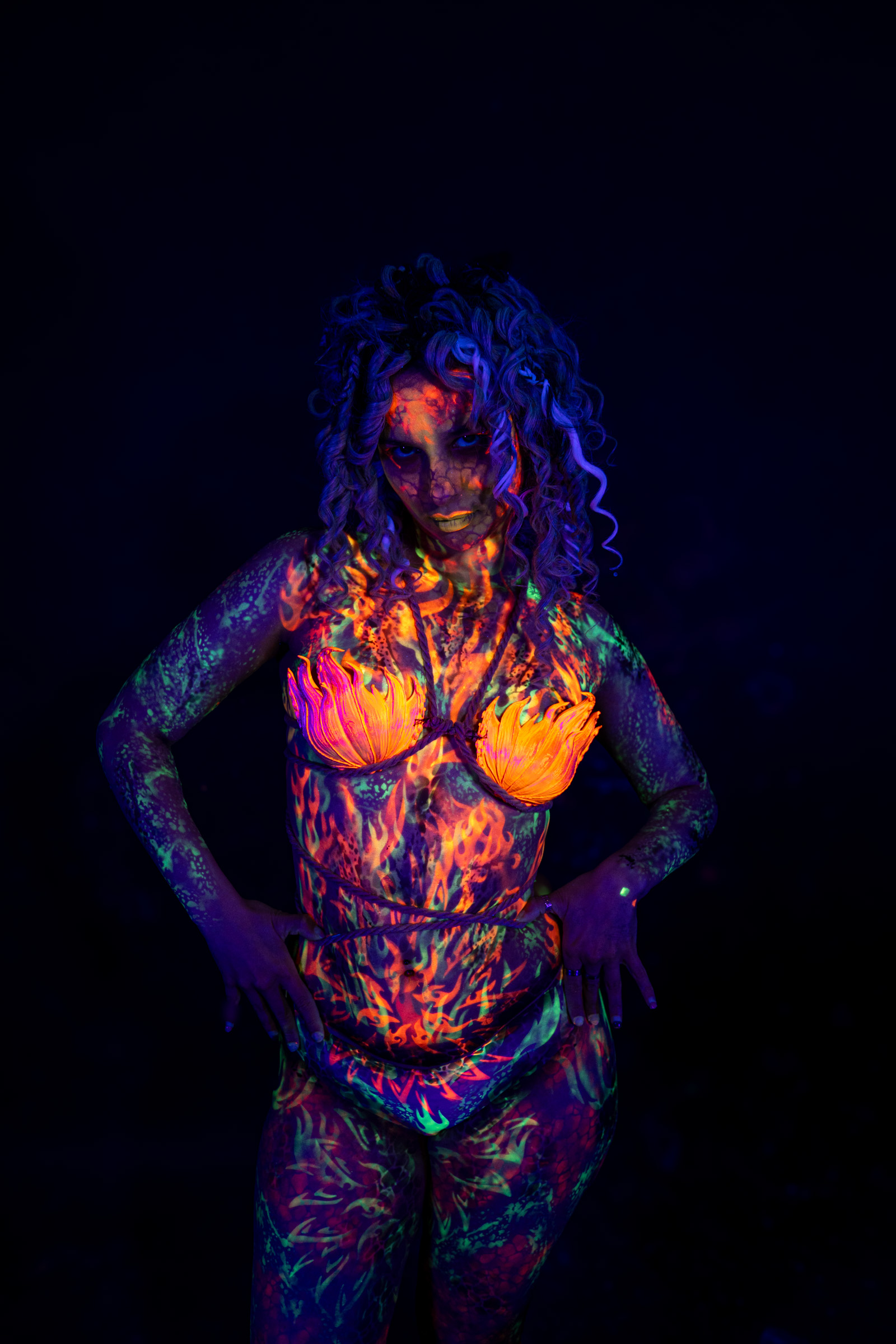 Bella stands facing the camera directly, curly hair framing her face. Orange, red, and green flame-patterned bodypaint covers her torso and limbs, with an orange flower-shaped design over the chest. Bodypaint by Elad Itzkin. Photographed by Elad Itzkin Photography under UV light in his studio in Gesundbrunnen, Berlin.