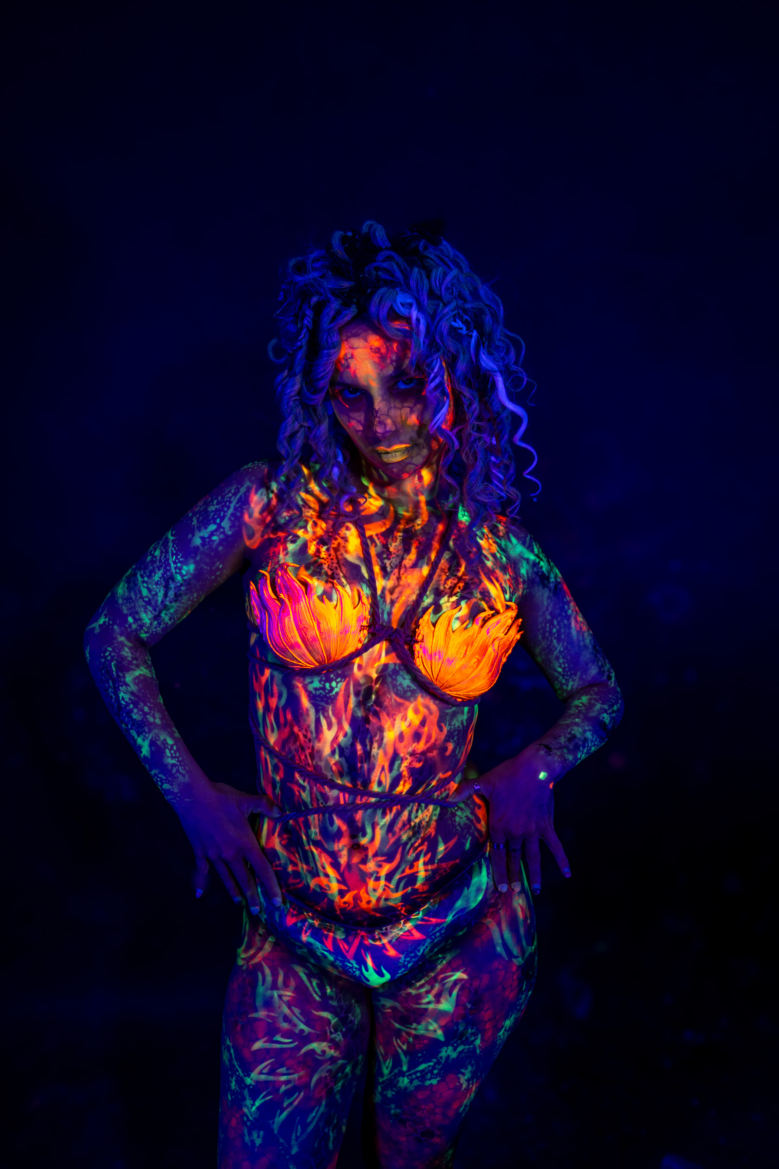 Bella stands facing forward in a direct pose. Her body is covered in an orange and red flame-style bodypaint design with green accents, including a flower-shaped motif over the chest. Bodypaint by Elad Itzkin. Photographed by Elad Itzkin Photography under UV light in his studio in Gesundbrunnen, Berlin.