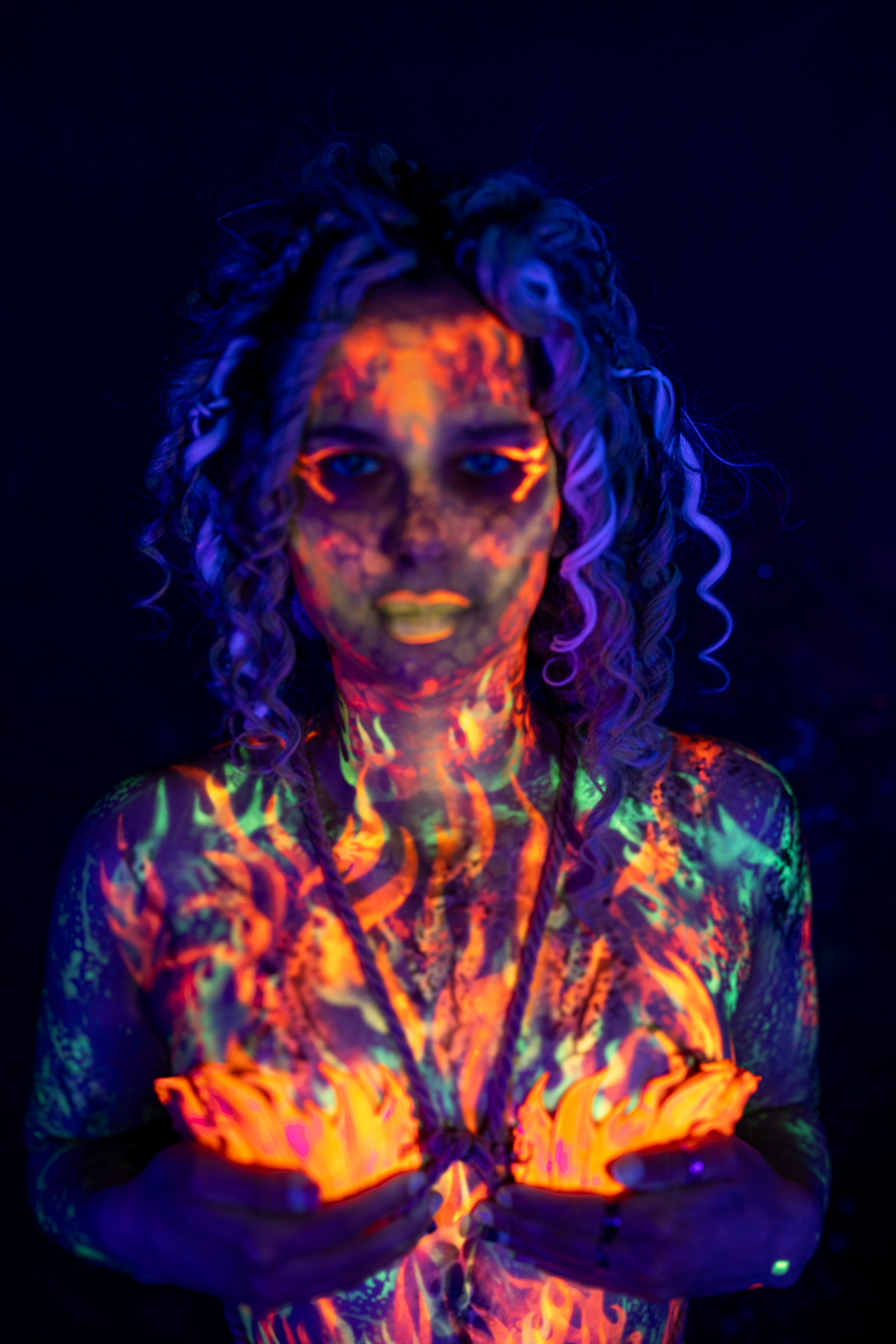Bella is shown from the torso up, standing and facing the camera. The design combines orange and red flame patterns across the face and body with green linework on the shoulders and chest. Bodypaint by Elad Itzkin. Photographed by Elad Itzkin Photography under UV light in his studio in Gesundbrunnen, Berlin.